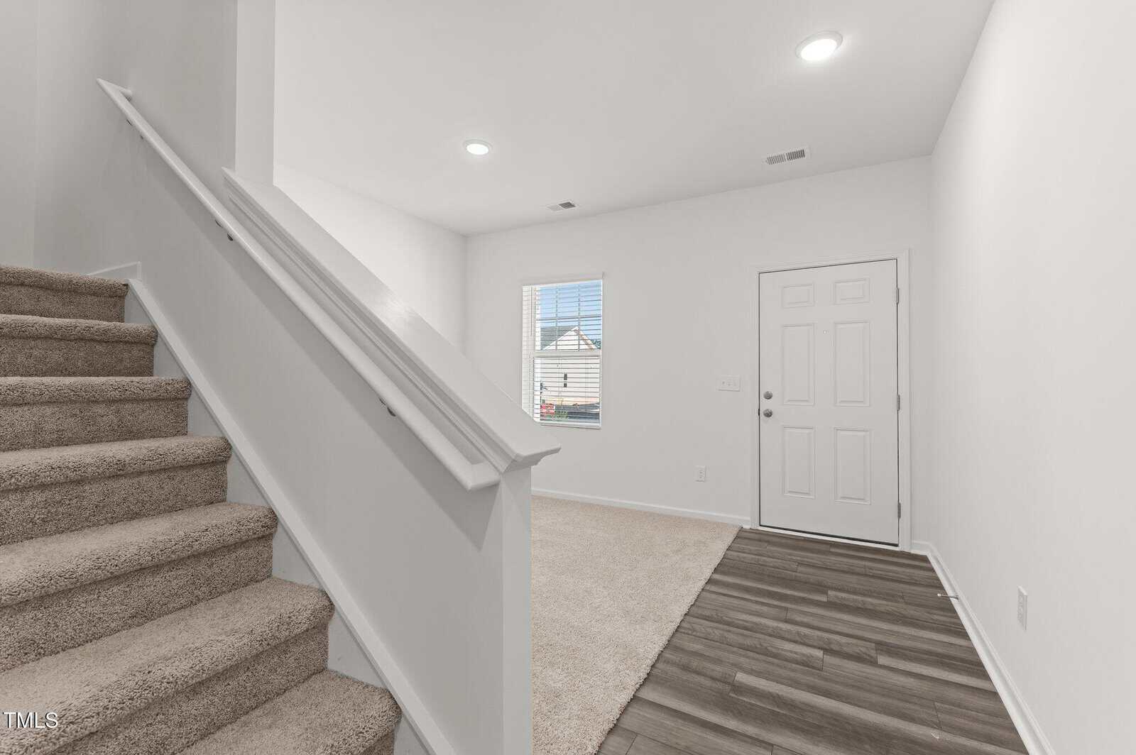 A view of a home entryway with a white front door, a window, carpeted stairs on the left, and a mix of carpet and wood flooring. The walls and ceiling are white with recessed lighting.