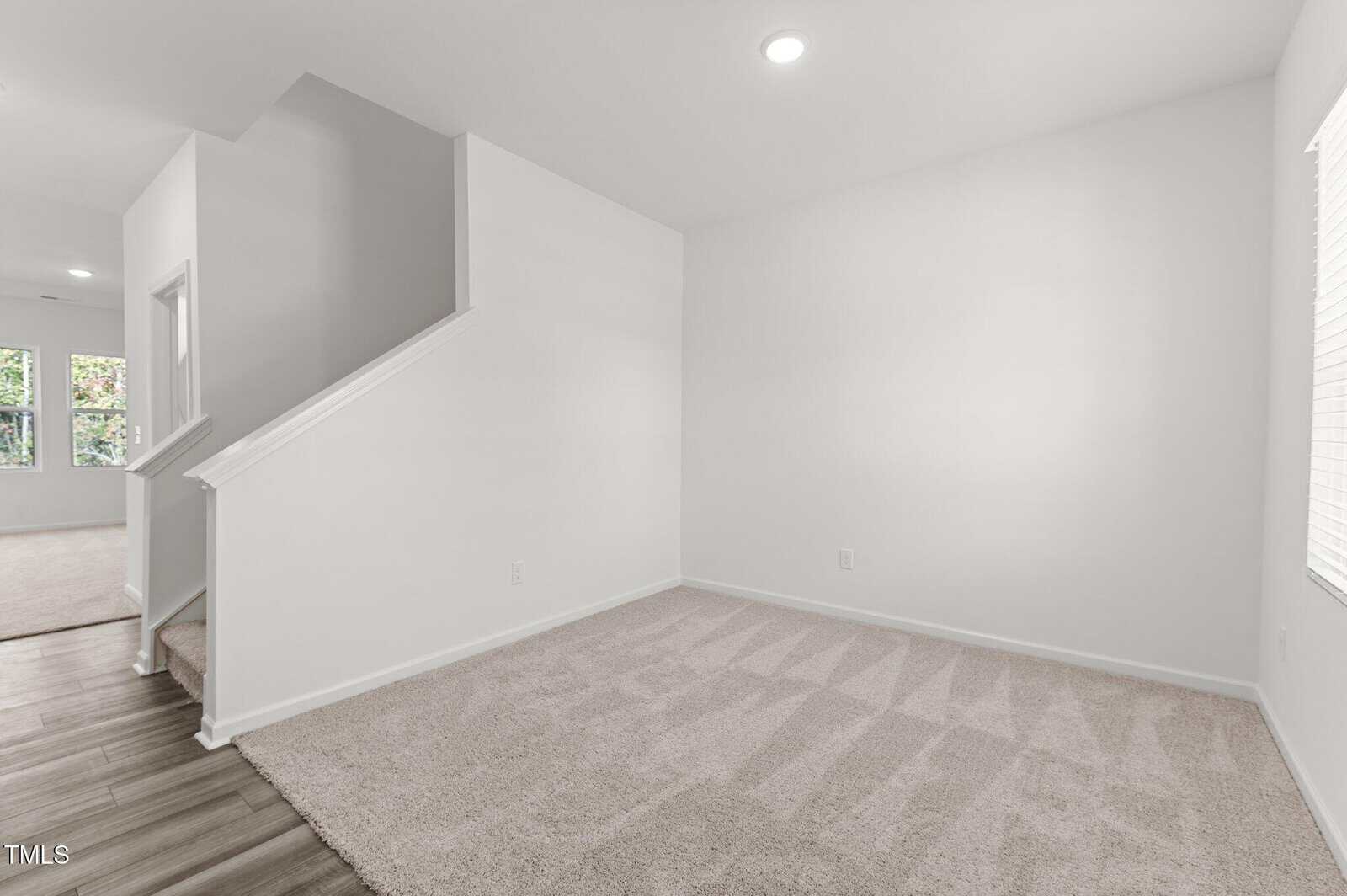 A clean, empty room with white walls, beige carpet, a staircase with a white railing, and a window with closed blinds. The floor transitions from carpet to wood at the base of the stairs.