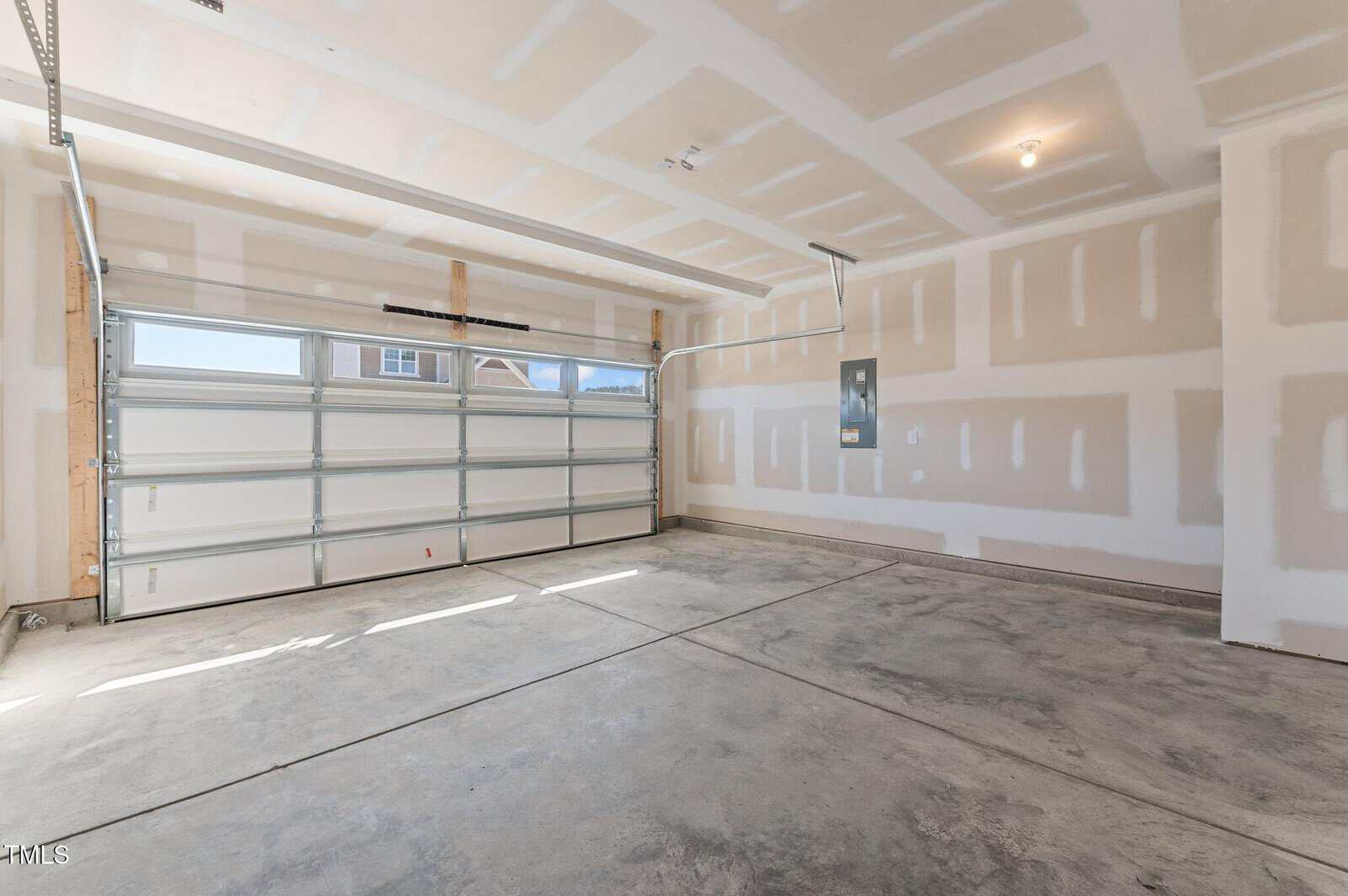 A clean, empty two-car garage with unfinished drywall, a concrete floor, an overhead garage door with windows, a ceiling light, and an electrical panel on the right wall.