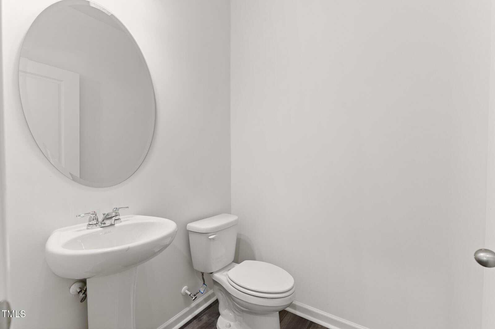 A small, white bathroom with a round mirror above a pedestal sink, a toilet next to it, and plain white walls.