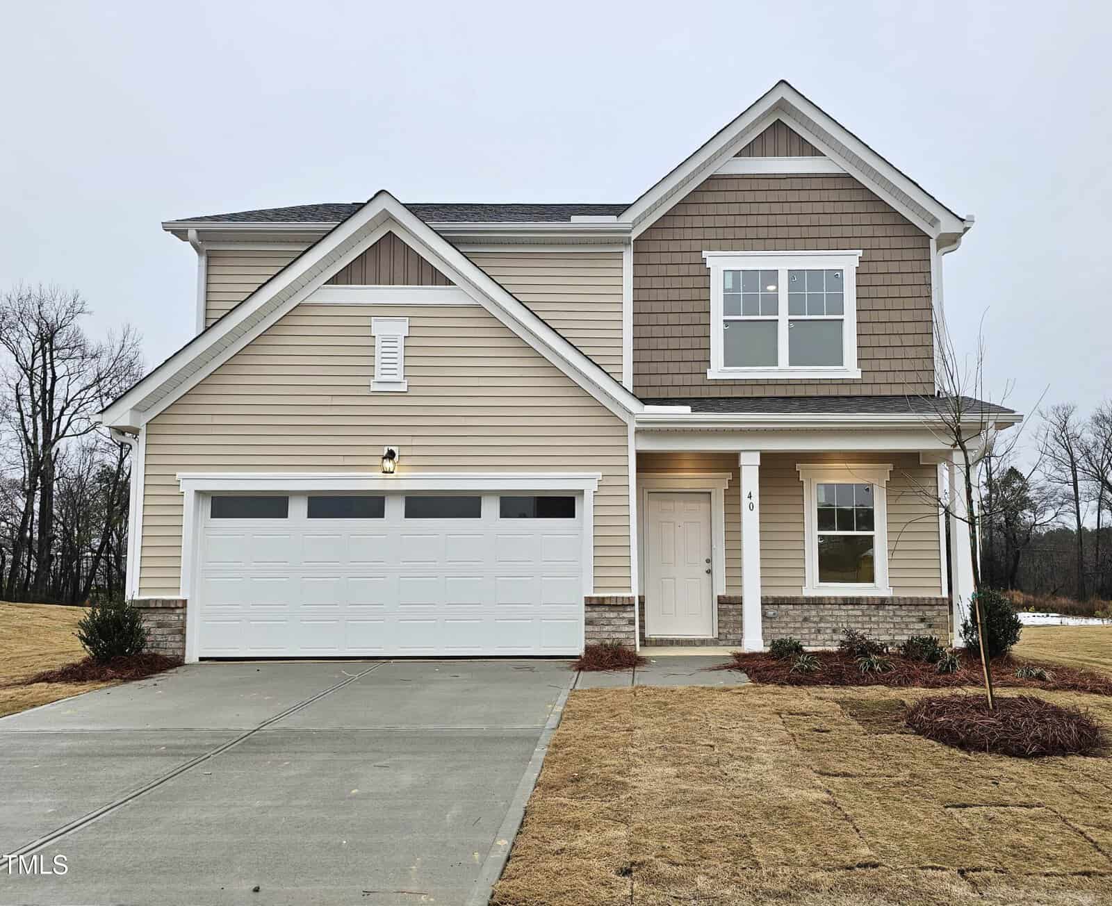 Two-story beige house with a double garage, white trim, and brown shingles on the upper level. The house has a small covered entryway, a concrete driveway, and a lawn with minimal landscaping.