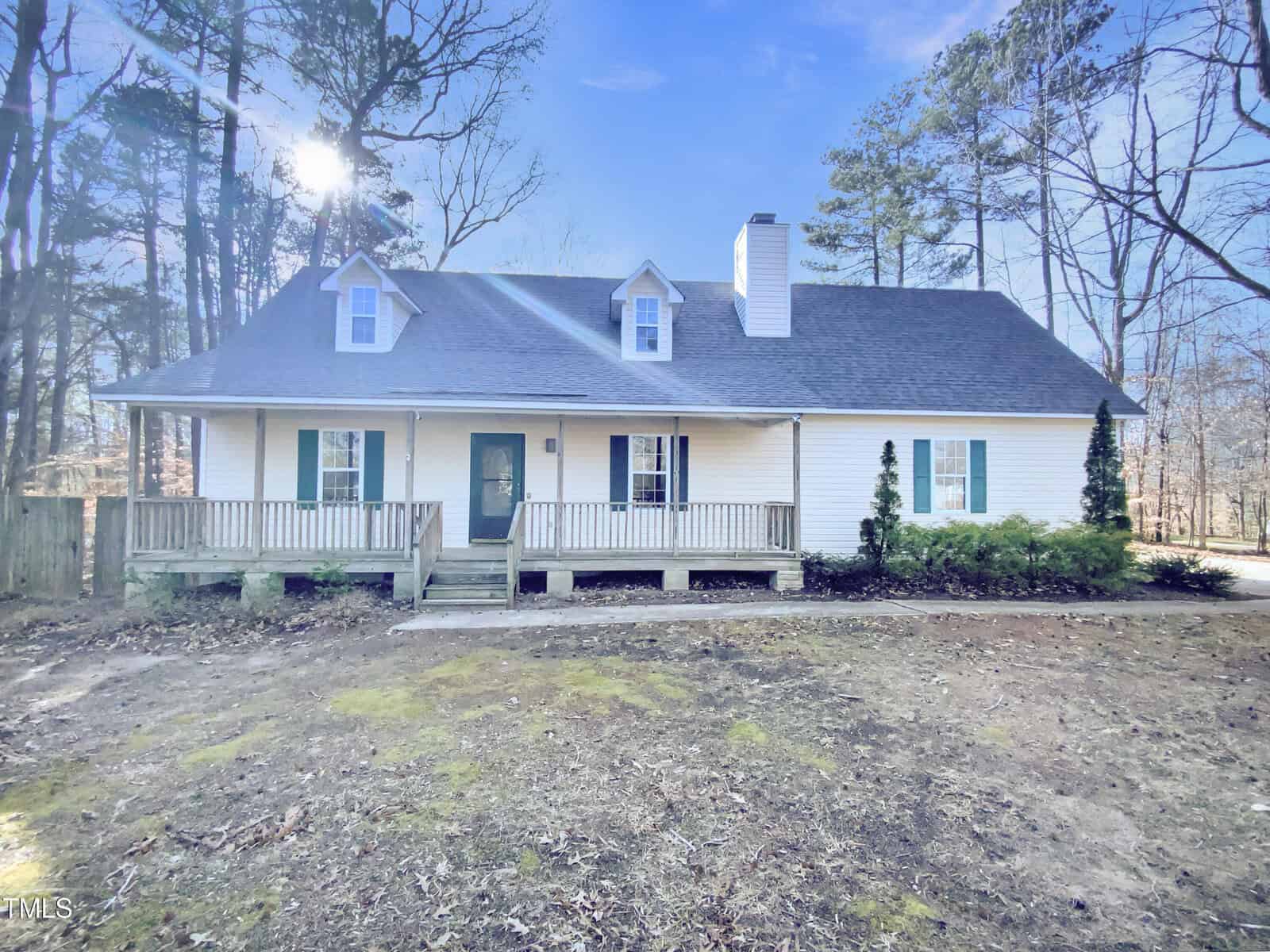 A white, one-and-a-half story house with a covered front porch, green door, dormer windows, and a shingled roof, surrounded by trees and a patchy lawn under a clear sky.