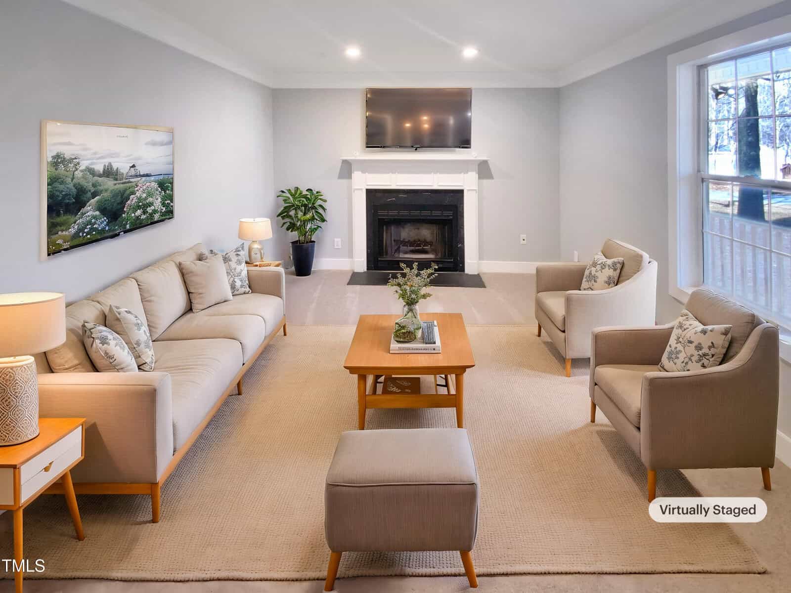 A living room with beige furniture, a sofa, two armchairs, a coffee table, and an ottoman on a light carpet. A TV is mounted above a fireplace, with a window on the right and a landscape painting on the left wall.