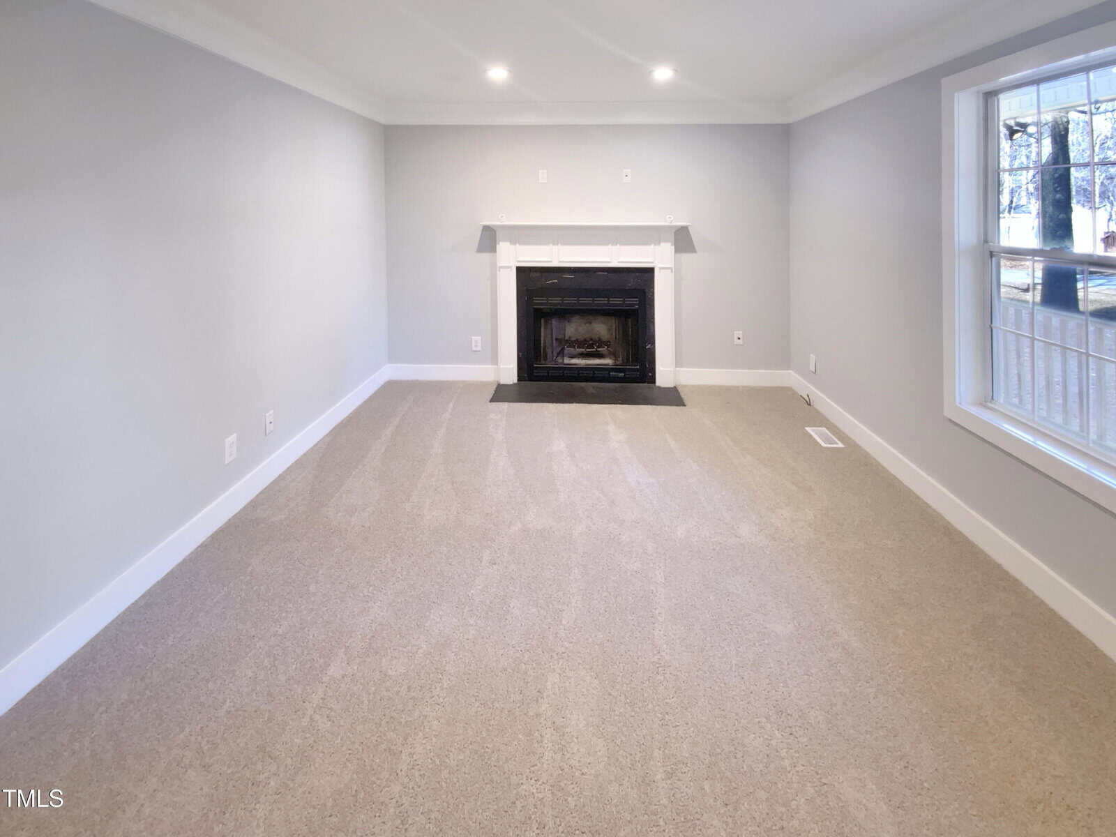 A carpeted living room with light gray walls, a white fireplace centered on the far wall, recessed ceiling lights, and a large window on the right side letting in natural light.