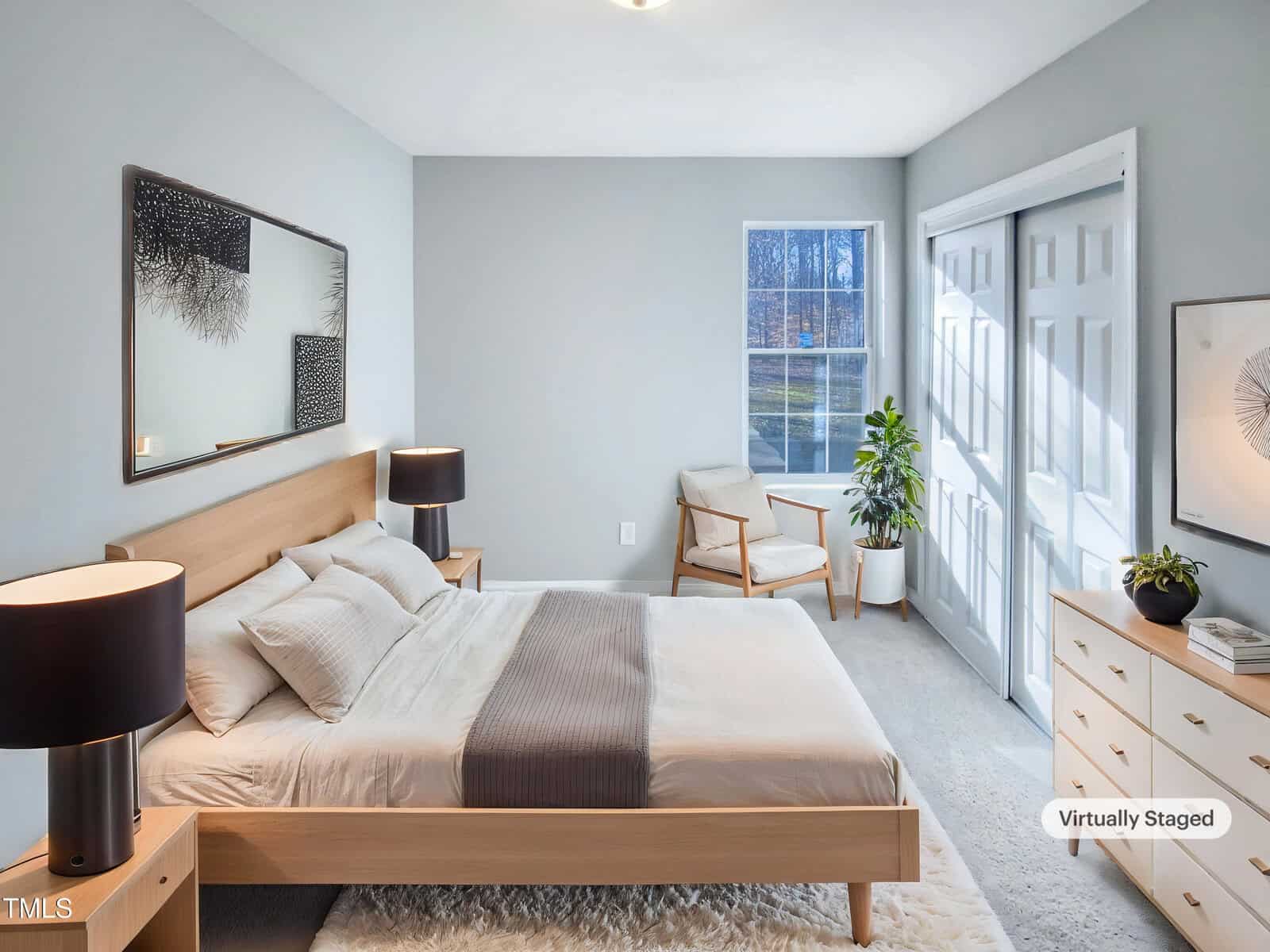 A modern bedroom with a wooden bed, white bedding, matching nightstands with black lamps, a dresser, a chair by the window, a potted plant, and framed wall art. The image is labeled "Virtually Staged.