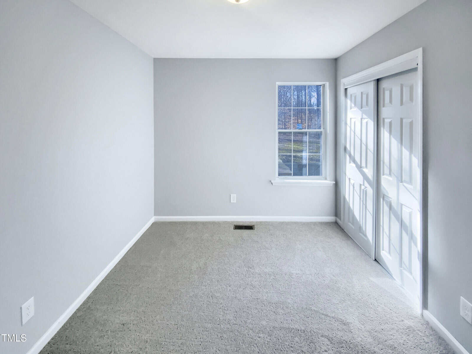 A small, empty room with light gray walls, beige carpet, a single window, and closed double closet doors. Sunlight is coming through the window, brightening the space.
