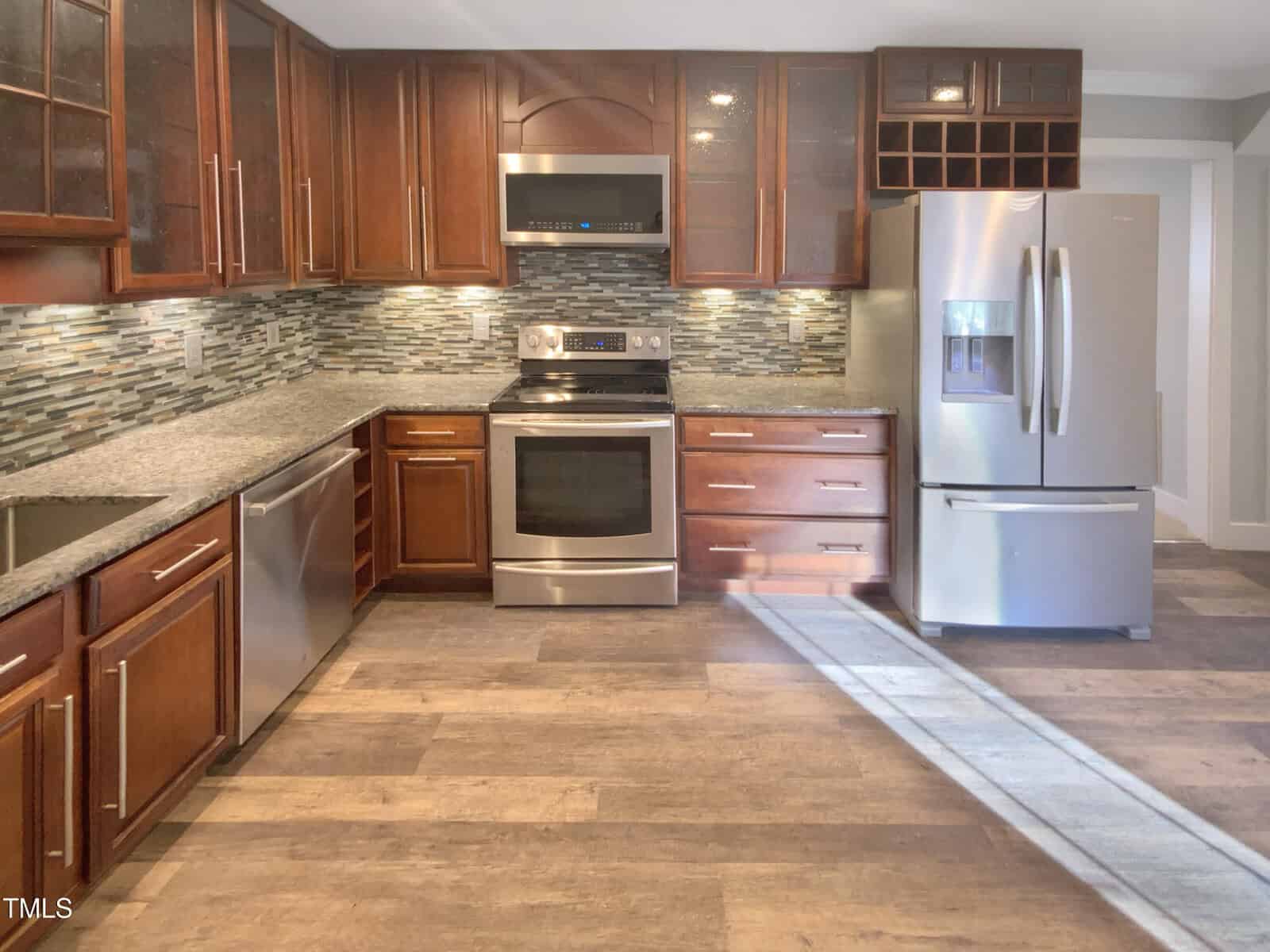 Modern kitchen with stainless steel appliances, dark wood cabinets, granite countertops, glass tile backsplash, and light wood flooring; sunlight shines in from the right.