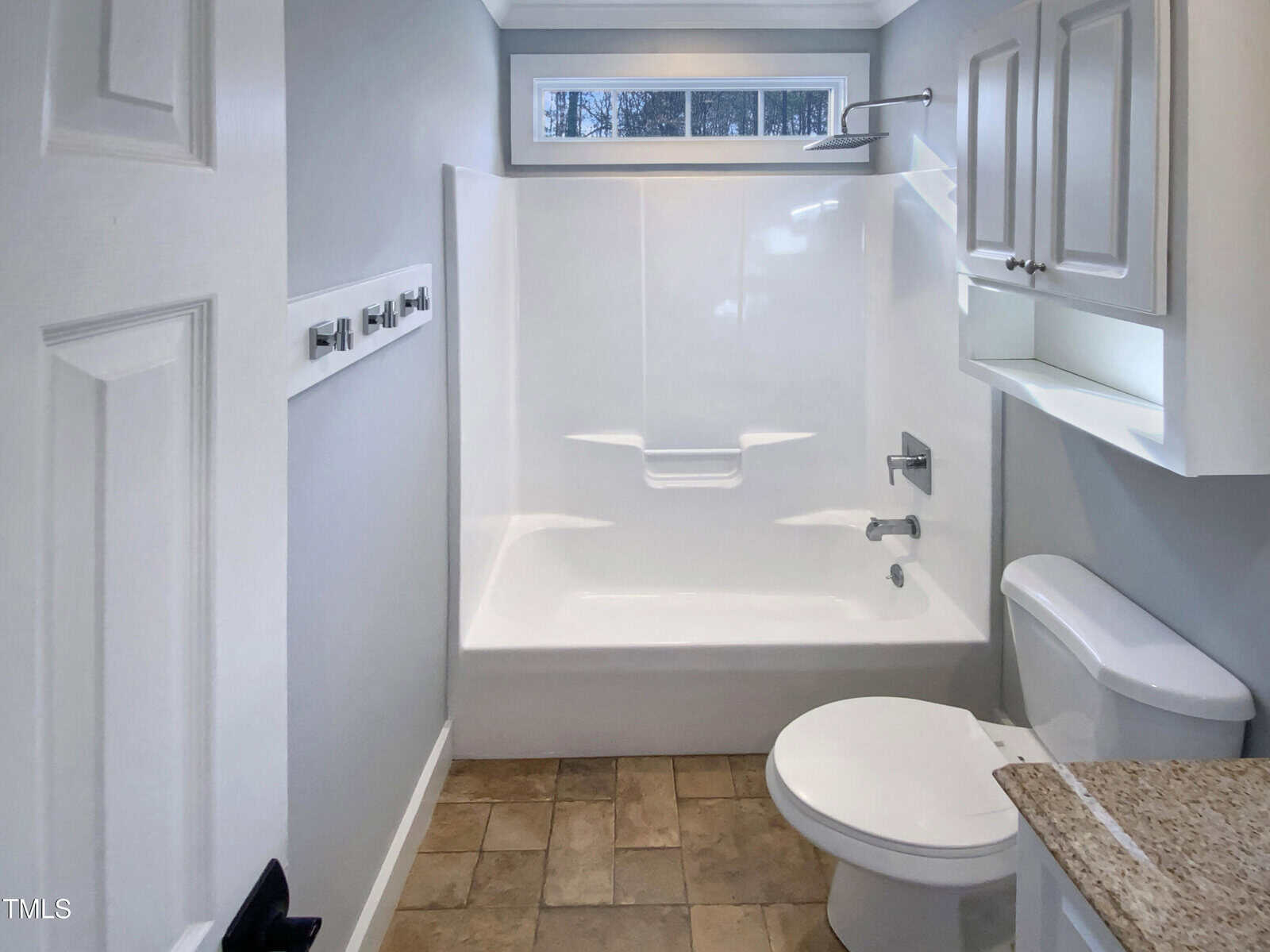 Bathroom with a white bathtub-shower combo, toilet, and sink with granite countertop. There are white cabinets above the toilet and neutral tile flooring. A small window is above the shower, letting in natural light.