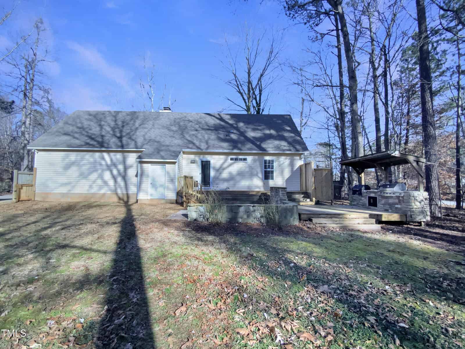 Single-story house with light siding and a gray roof, pictured from the backyard. A wooden deck with a covered hot tub is on the right, surrounded by trees and winter foliage. Shadows from trees stretch across the yard.