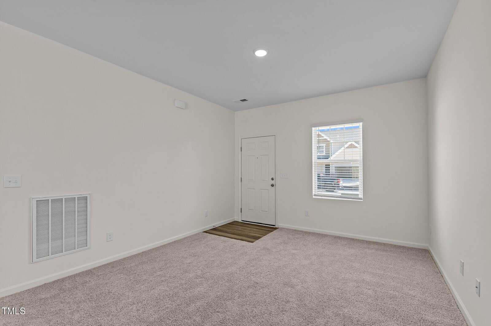 Empty room with light beige walls, carpeted floor, and a single window letting in natural light. There’s a closed white door near the window and a small vent on the left wall.