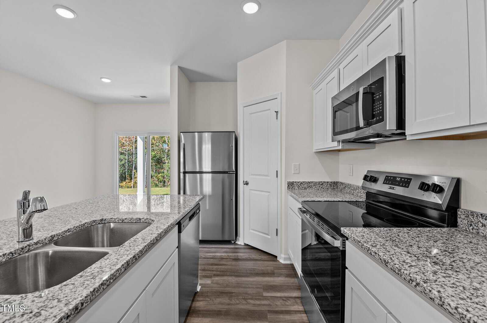 Modern kitchen with white cabinets, granite countertops, stainless steel appliances including a fridge, microwave, and stove, dual sink, and wood-style flooring. Glass doors in the background open to a grassy outdoor area.