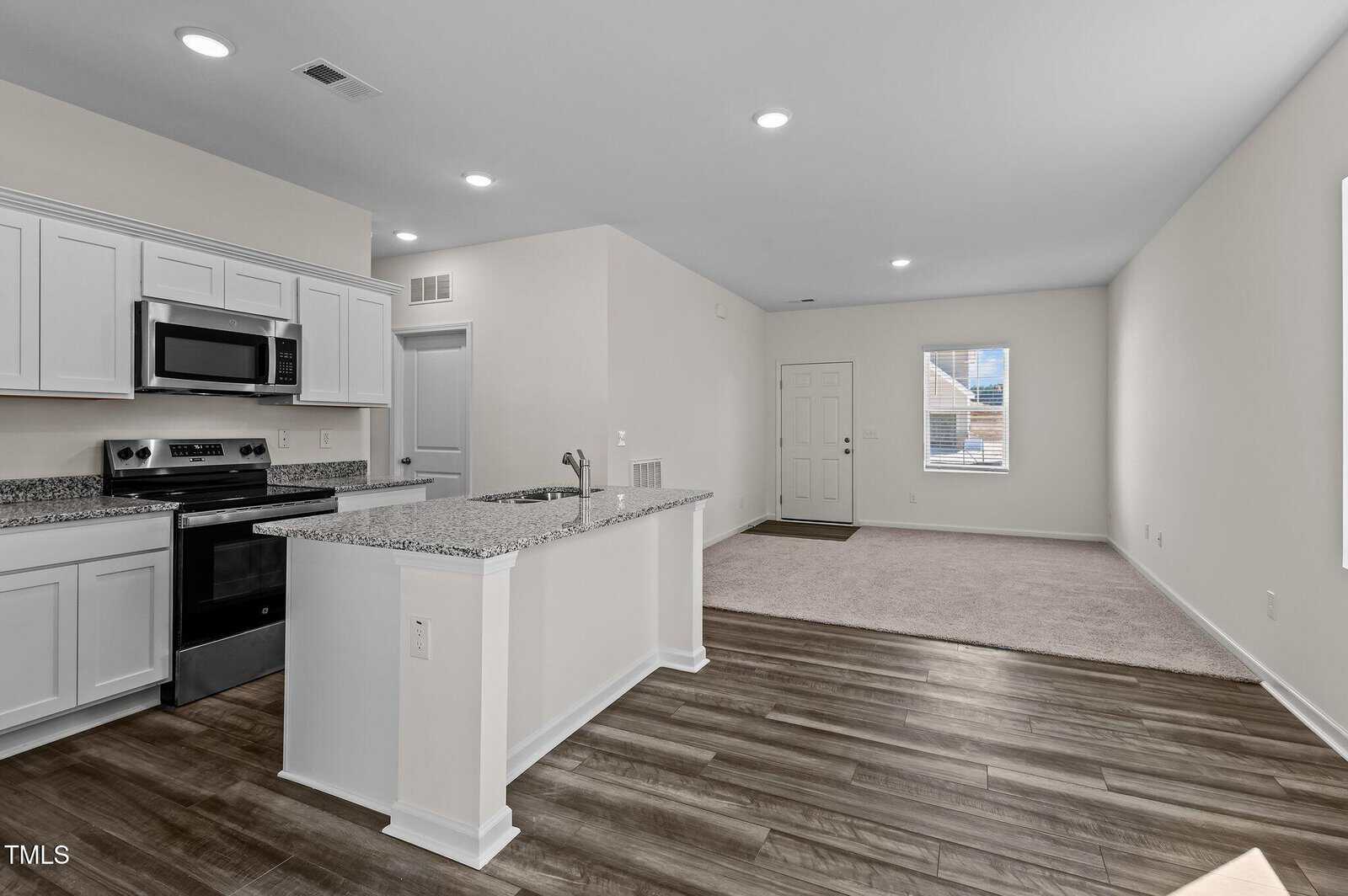 A modern open-concept kitchen and living area with white cabinets, stainless steel appliances, granite countertops, wood flooring in the kitchen, and carpet in the living room. A window and front door are visible in the background.