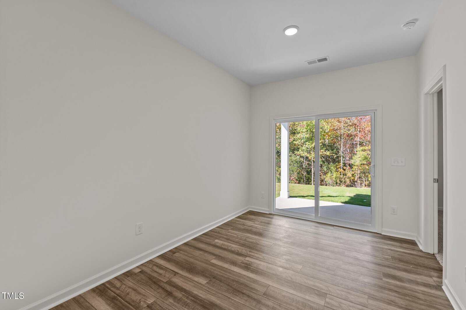 Empty room with light-colored walls, wood flooring, a ceiling light, and large glass doors leading to a patio and green outdoor area. There is also a small open doorway on the right side.