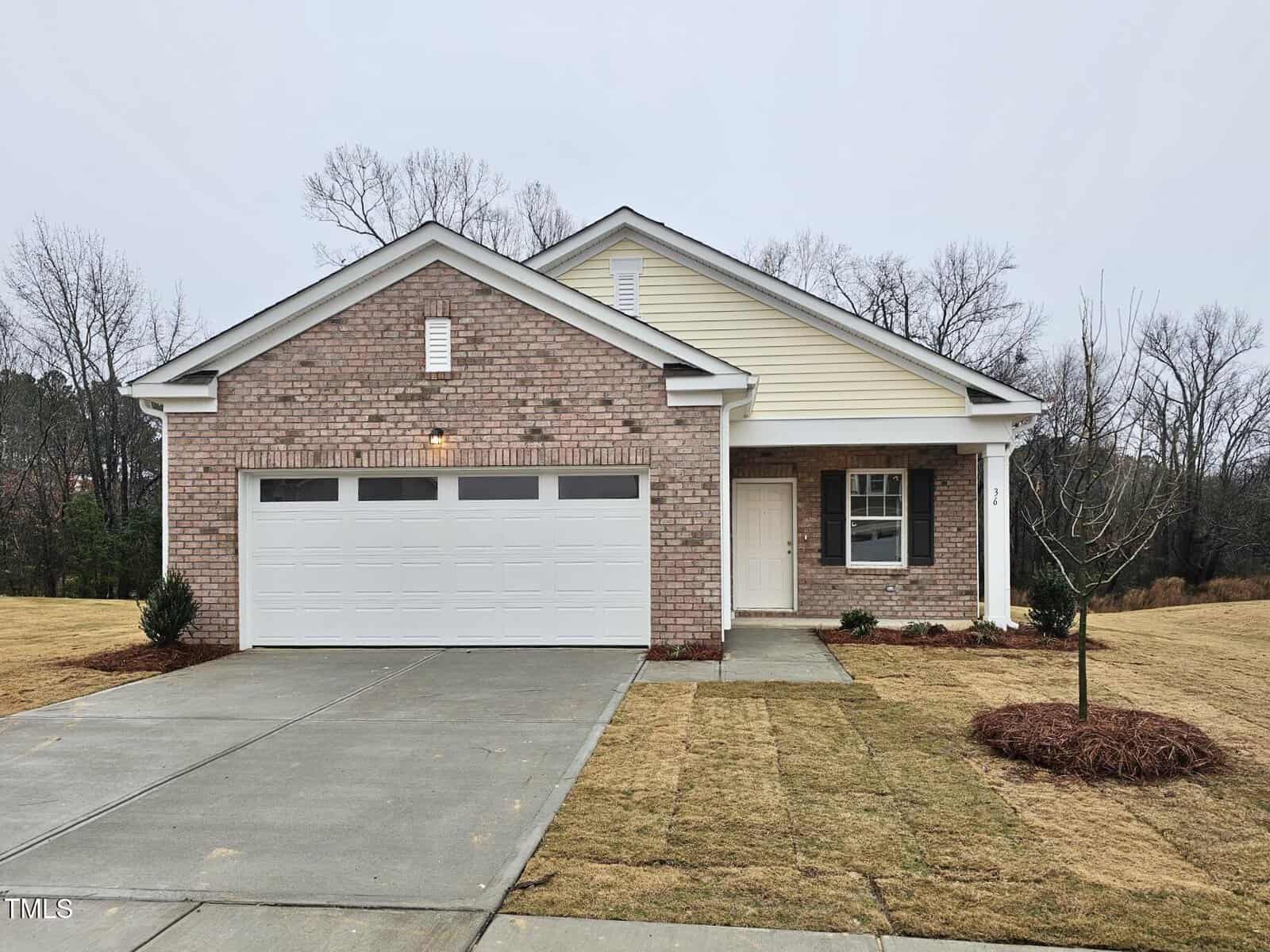 A single-story house with a brick and siding exterior, two-car garage, covered front porch, and a small tree planted in the front yard, set against a backdrop of bare trees and cloudy sky.