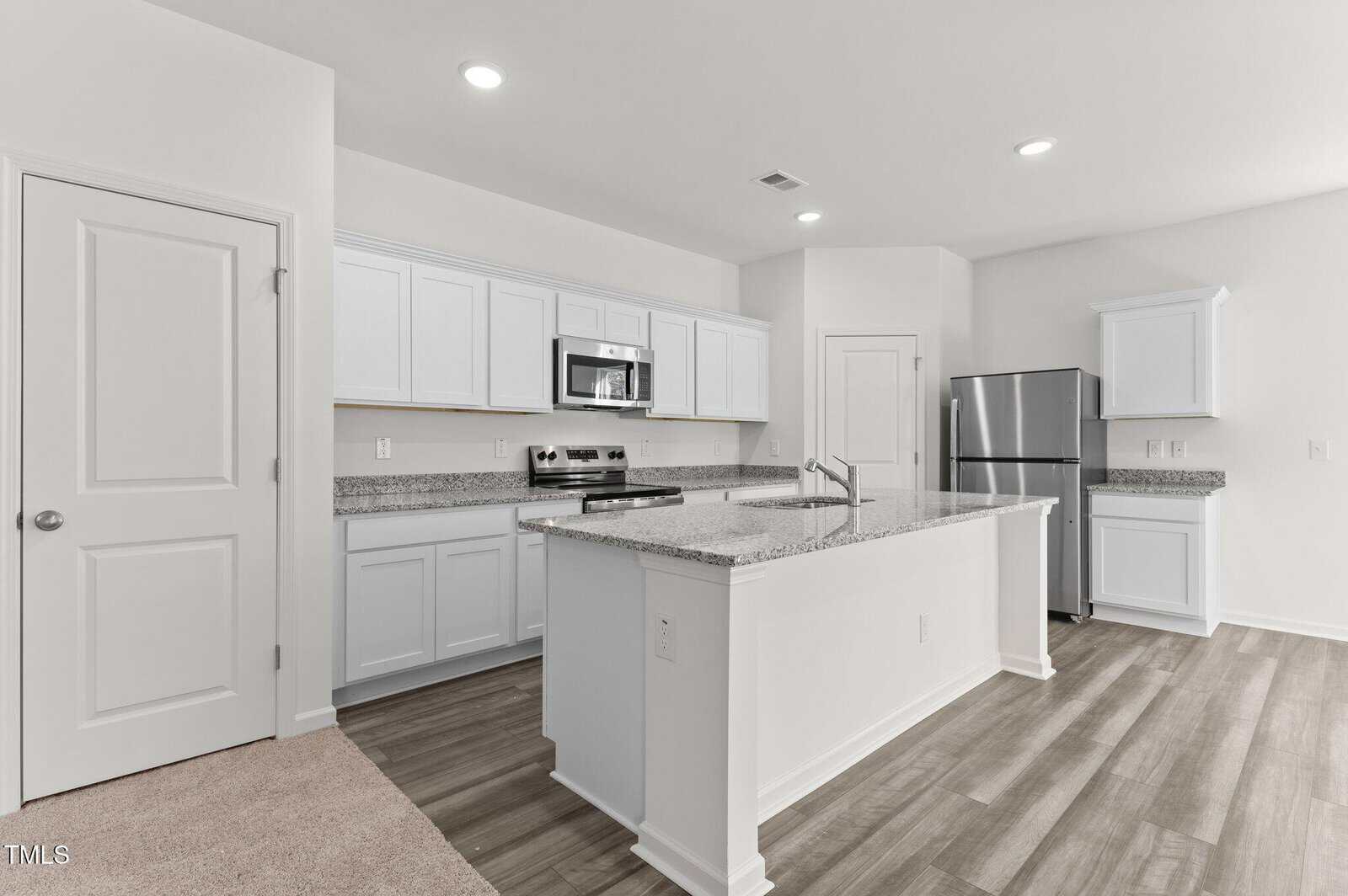 A modern kitchen with white cabinets, granite countertops, stainless steel appliances, an island with a sink, and wood-style flooring. Walls are white, and lighting is recessed in the ceiling.