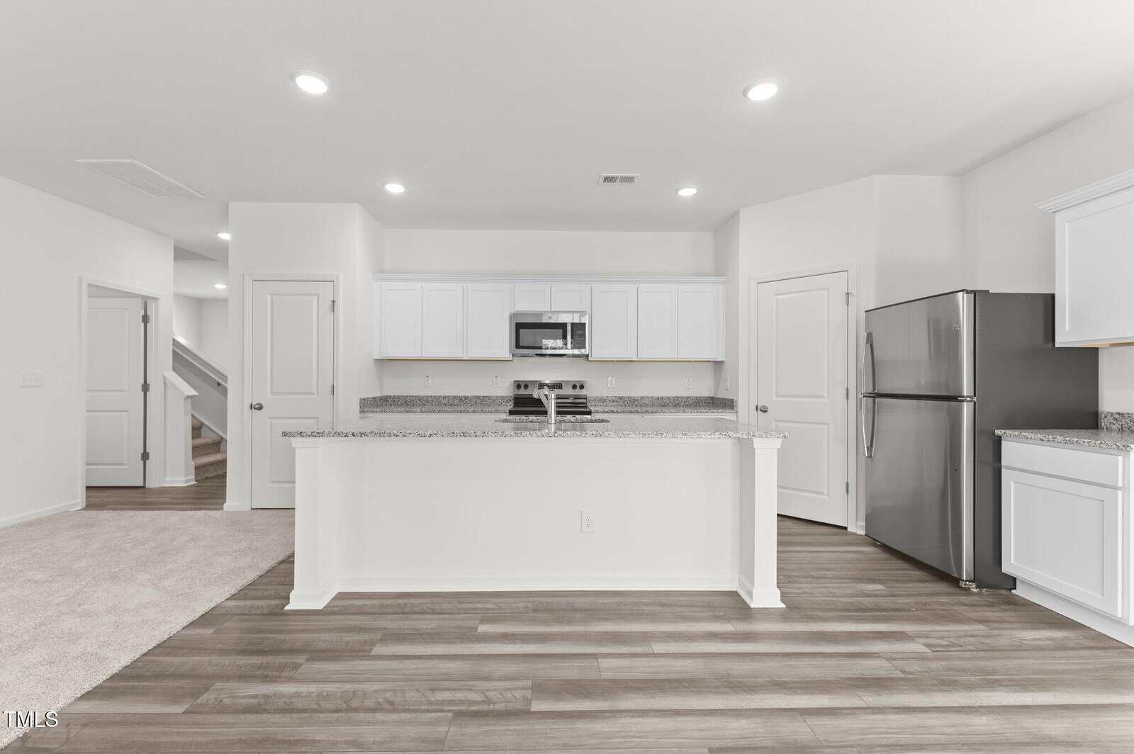 A modern kitchen with white cabinets, granite countertops, stainless steel refrigerator, and electric stove. The room has wood-style flooring and recessed lighting, with a carpeted area and staircase visible to the left.