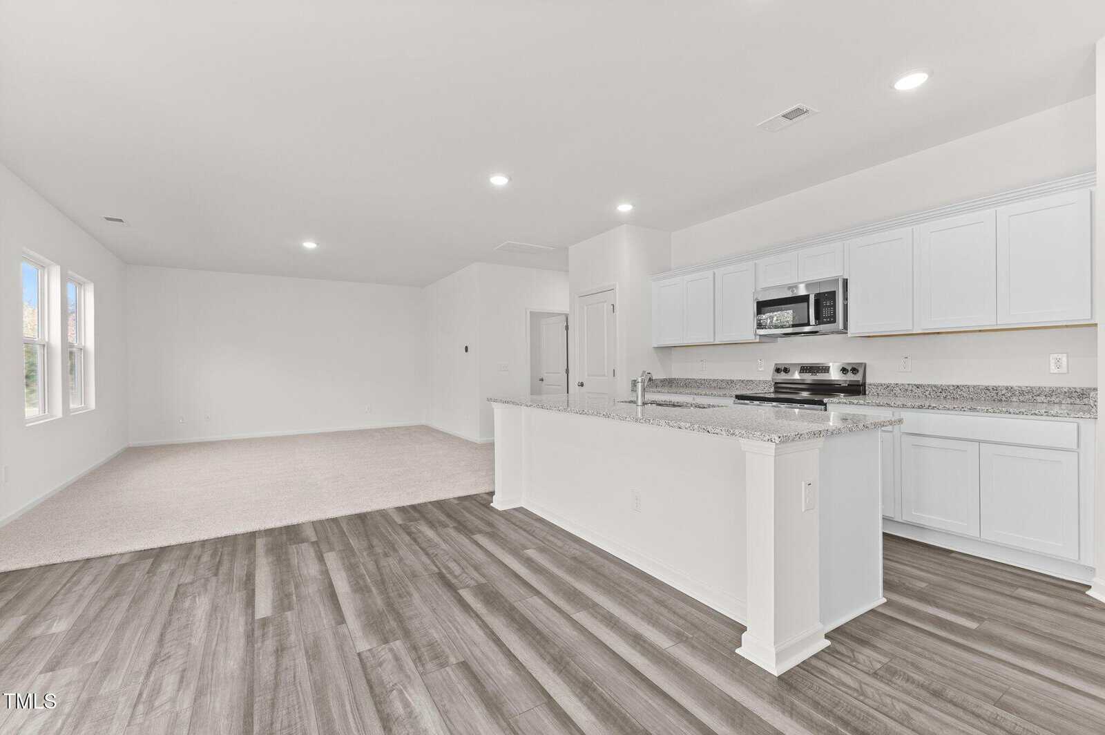 Open-concept living area with wood-look floors, adjoining kitchen with white cabinets, granite countertops, stainless steel appliances, and a carpeted area in the background with large windows.