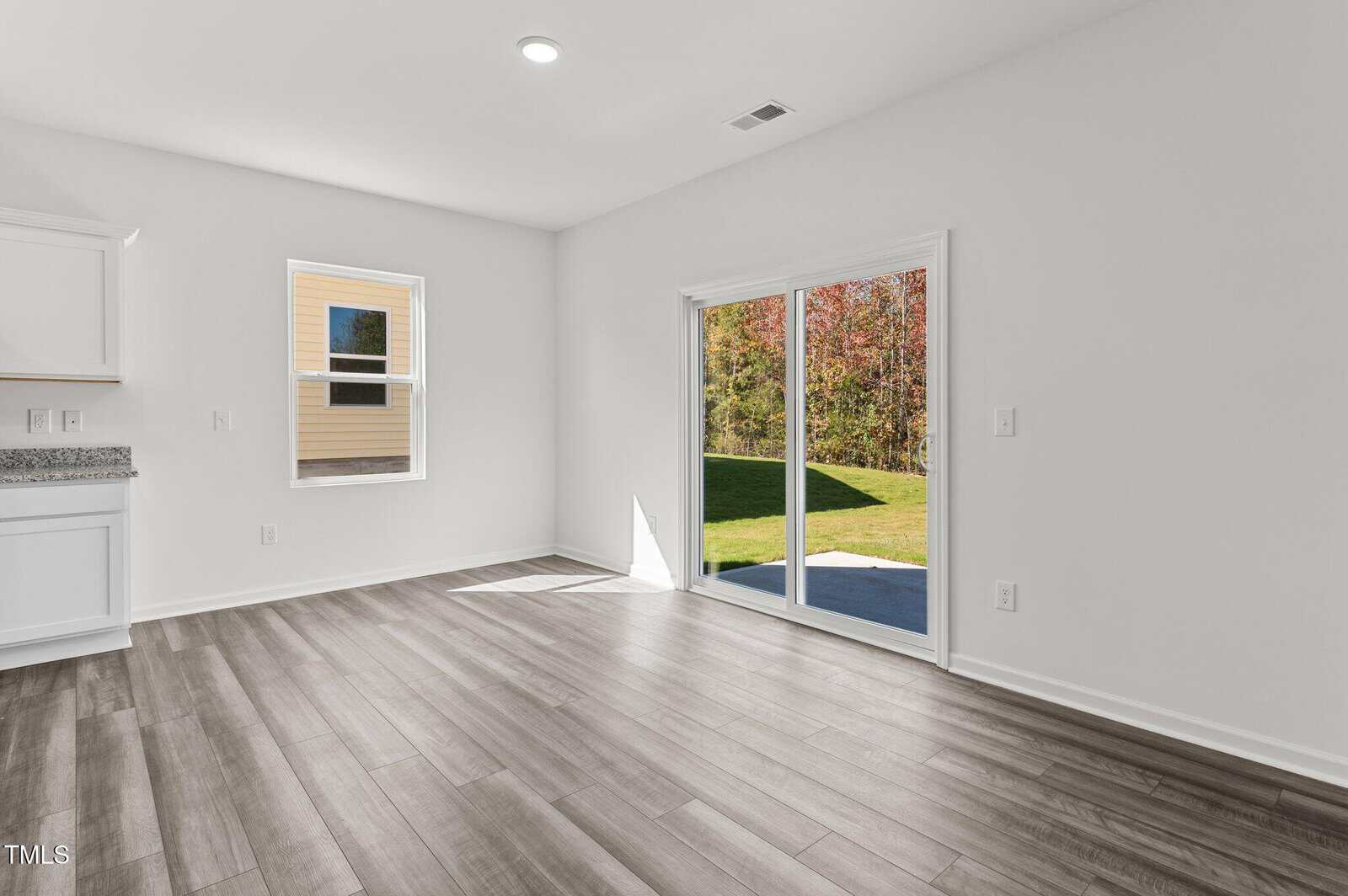 Bright, empty room with light wood flooring, white walls, and a sliding glass door leading to a green backyard; a single window lets in more sunlight.