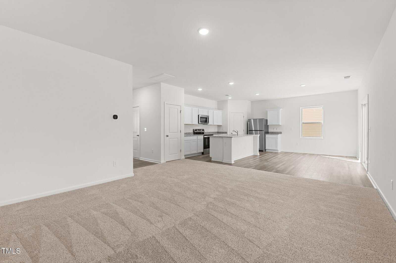 Spacious open-concept living area with beige carpet, white walls, and a modern kitchen featuring white cabinets, stainless steel appliances, and wood-look flooring. Large windows provide natural light.