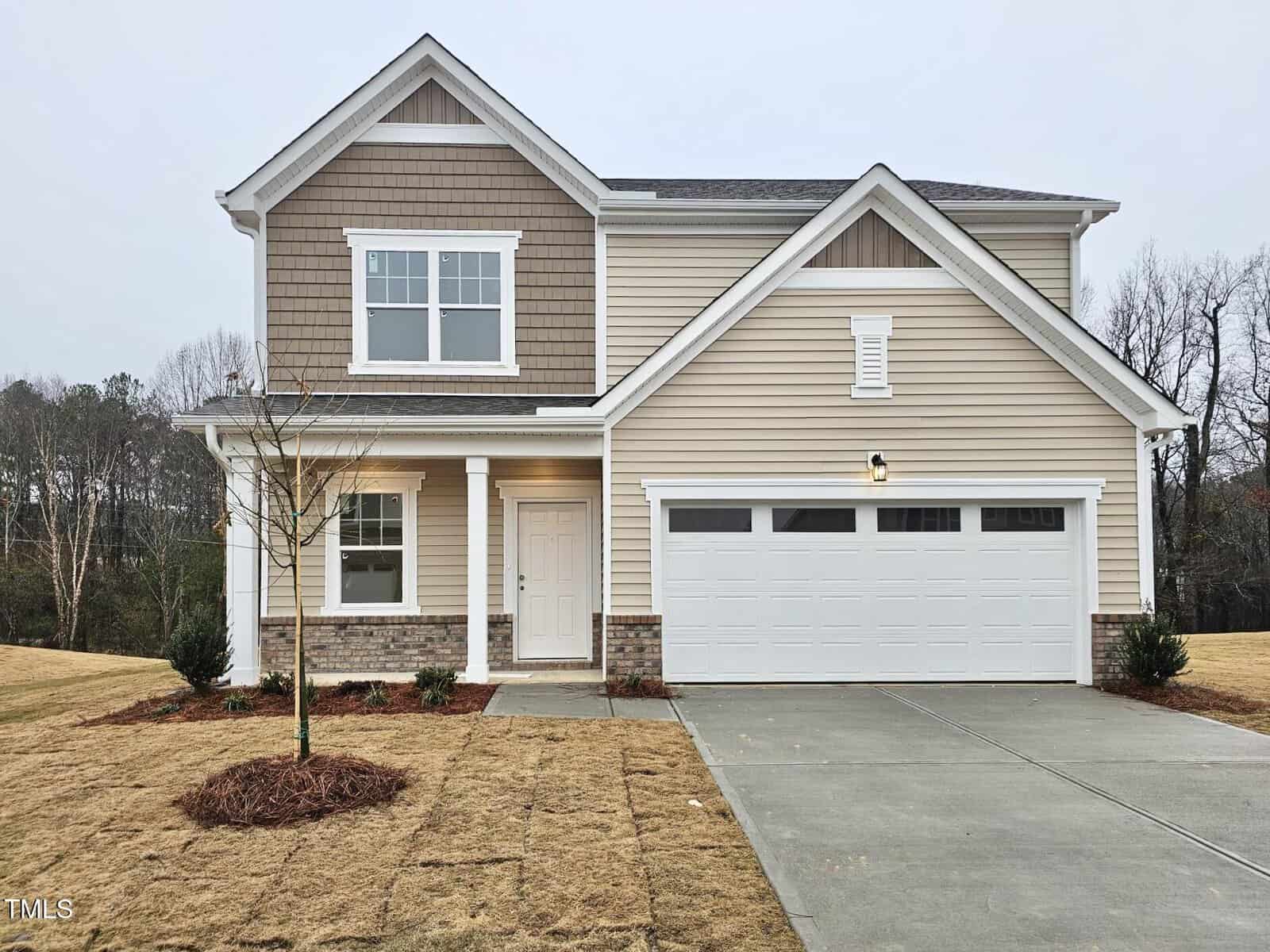 Two-story suburban house with beige siding, white trim, a covered front porch, and a double garage. A small tree and minimal landscaping are in the front yard, and the sky is overcast.