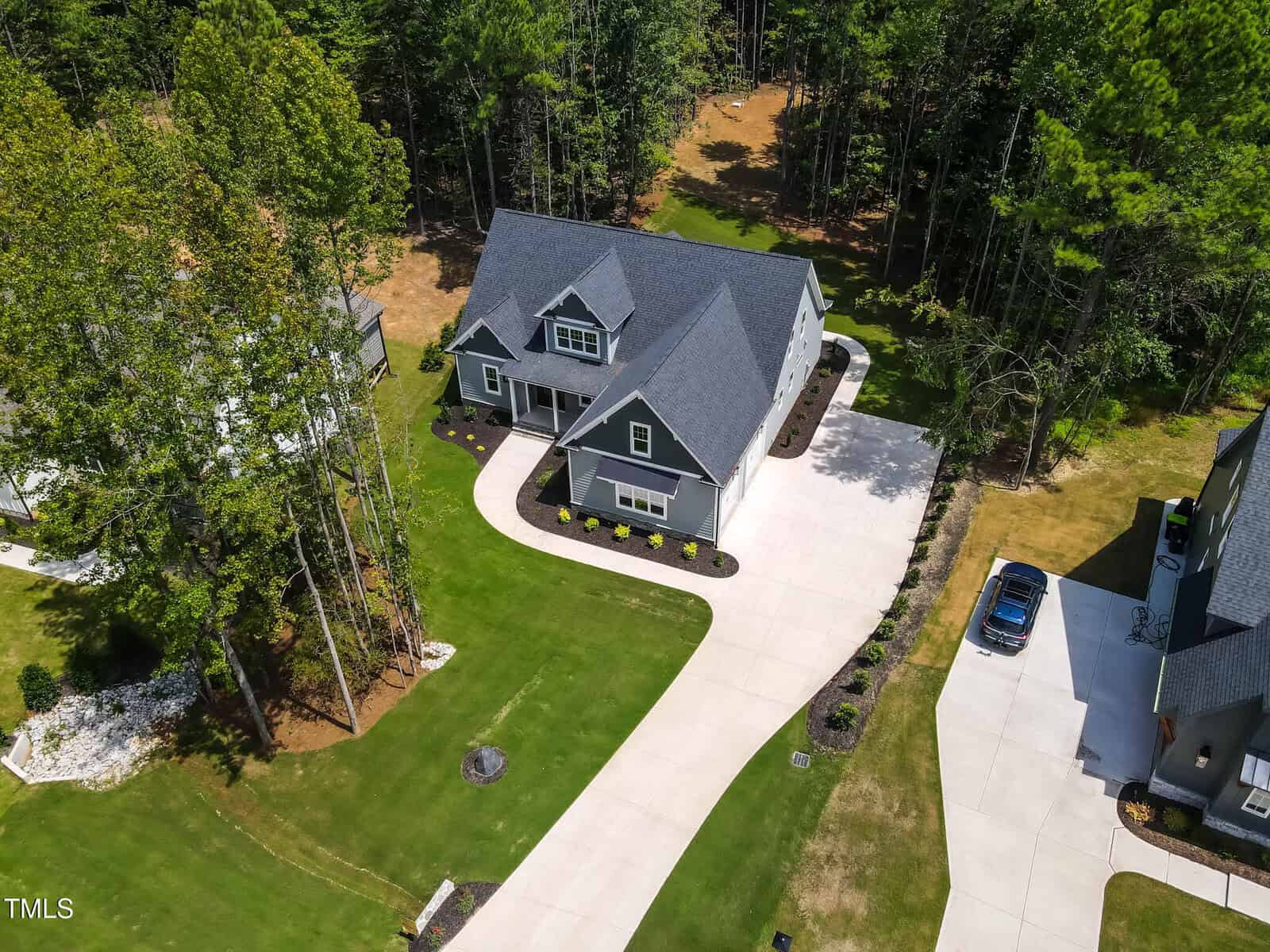 Aerial view of a two-story house with a dark roof, gray exterior, and white trim. The property has a large driveway, neatly manicured lawn, surrounding trees, and neighboring houses.