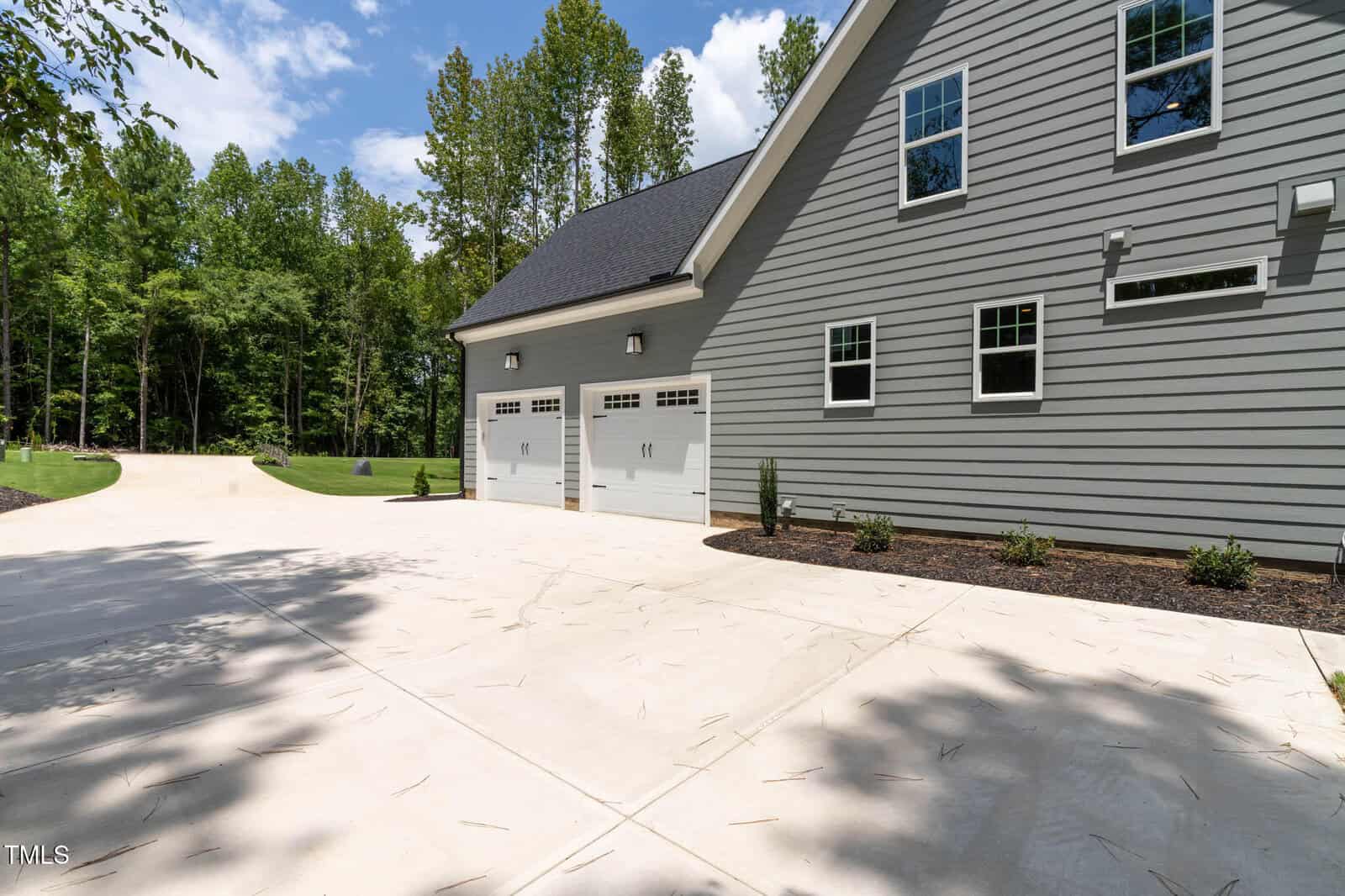The image shows a large concrete driveway leading to a three-car garage attached to a gray house with white trim, surrounded by trees and landscaped with small shrubs.