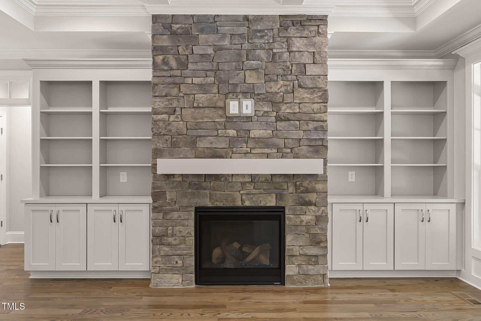 A stone fireplace with a white mantel is centered between two built-in white shelving units with cabinets below. The room has hardwood floors and white trim.