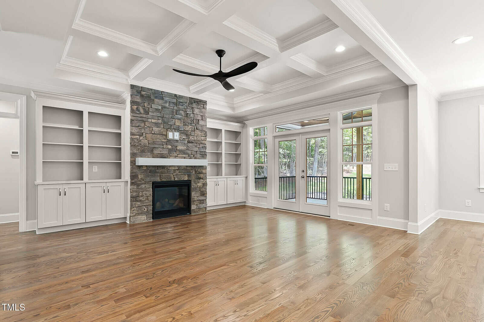 A spacious living room with wood floors, built-in white shelves, a stone fireplace, coffered ceiling, large windows, and glass doors opening to a covered patio with a view of a green yard.