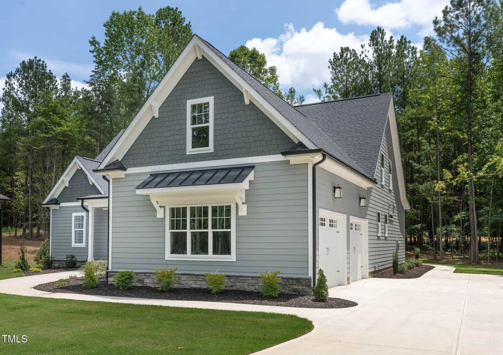 A modern, light gray house with white trim, a steep gabled roof, and a two-car garage sits on a concrete driveway surrounded by trees and neatly landscaped shrubs.