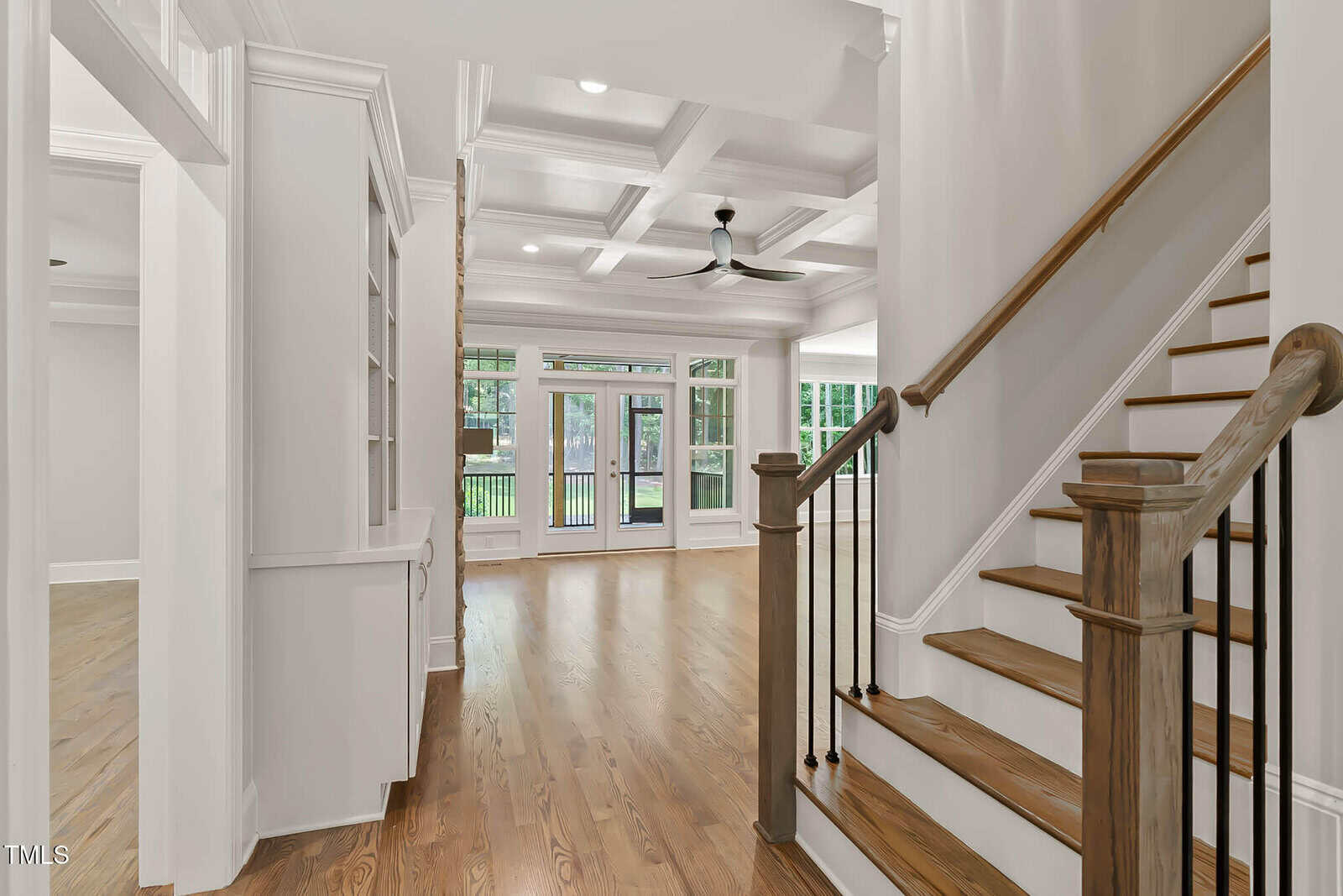 A view of a modern home interior featuring a wooden staircase with white risers and black balusters, coffered ceilings, hardwood floors, built-in shelves, and large windows overlooking a backyard.