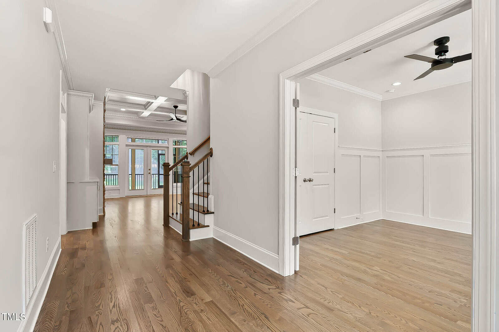 A hallway with hardwood floors leads to a living area with large windows and glass doors. On the right, an open doorway reveals a room with wainscoting and a ceiling fan. A staircase is visible in the center.