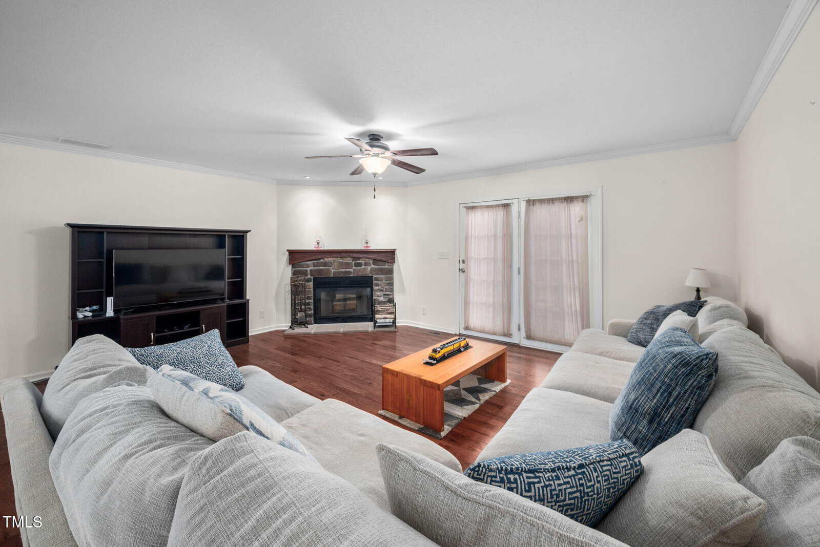 Living room with a large beige sectional sofa around a wooden coffee table. A stone fireplace sits against the far wall. A ceiling fan is overhead, and a TV on a dark stand is on the left. Double doors with blinds are on the right side.