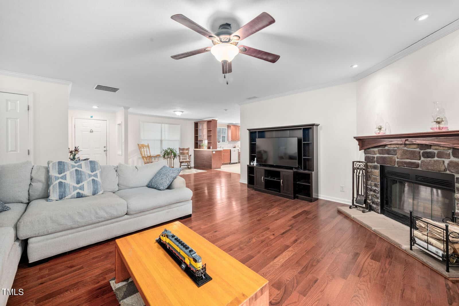 A living room with hardwood floors features a light gray sectional sofa, a wooden coffee table with a toy train, a flat-screen TV on a black stand, and a stone fireplace. The space is well-lit with recessed lighting and a ceiling fan.