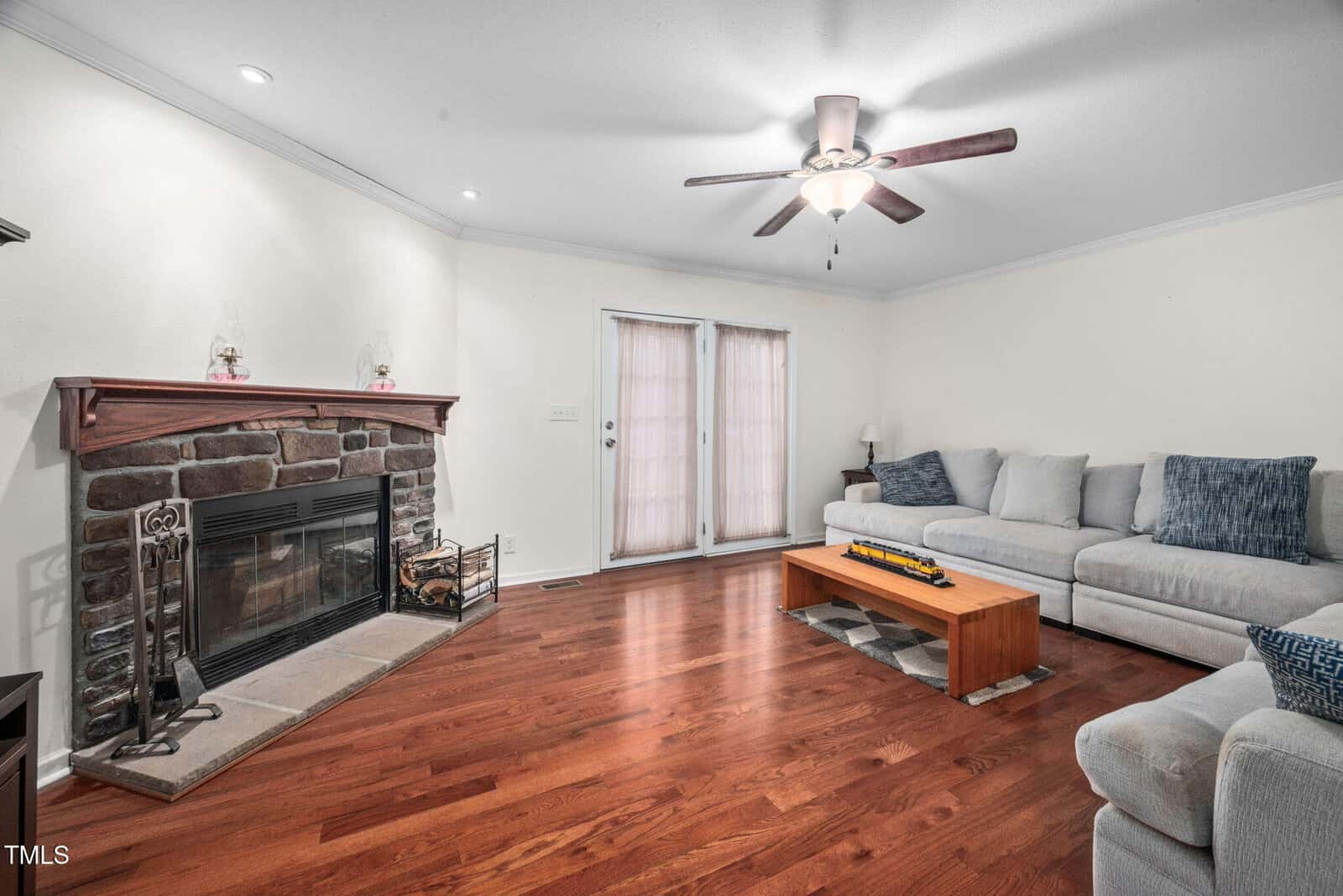 A living room with wooden flooring, a stone fireplace, and a ceiling fan. A large L-shaped sofa with gray cushions is against the wall. A wooden coffee table is in front of the sofa. French doors with curtains are in the background.
