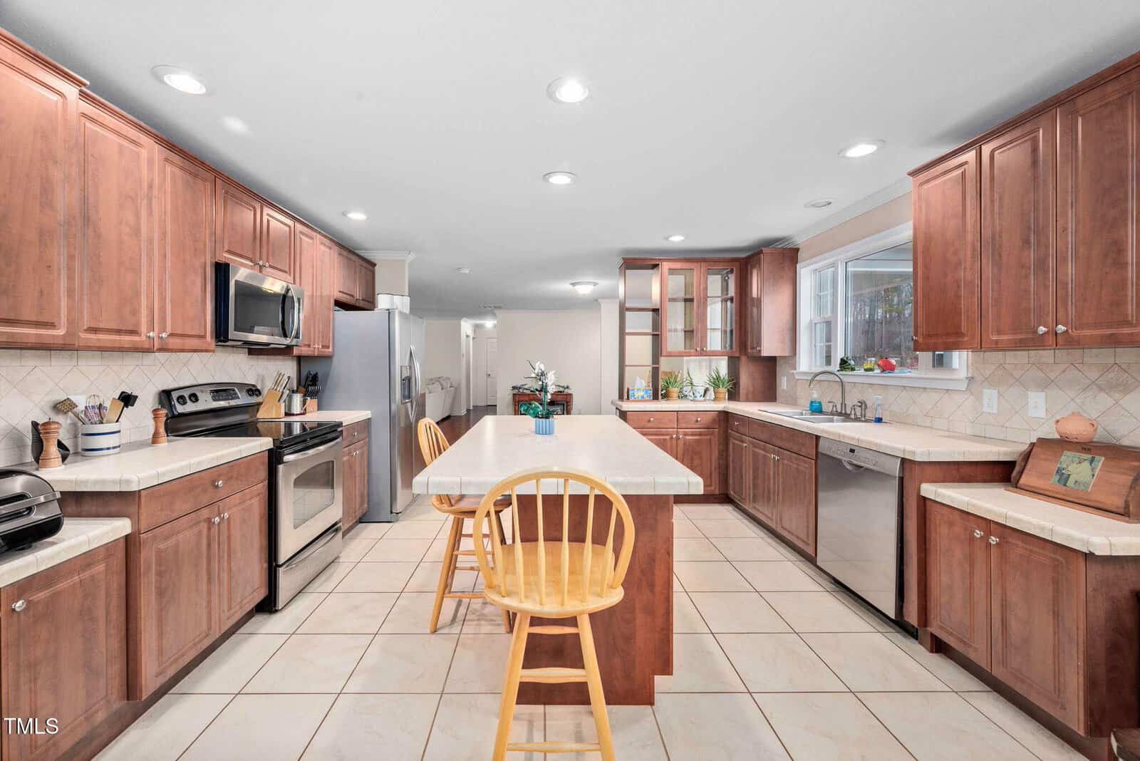 A spacious kitchen with wooden cabinets, stainless steel appliances, and a large central island with bar stools. The room features tiled floors, a window above the sink, and a decorative backsplash.