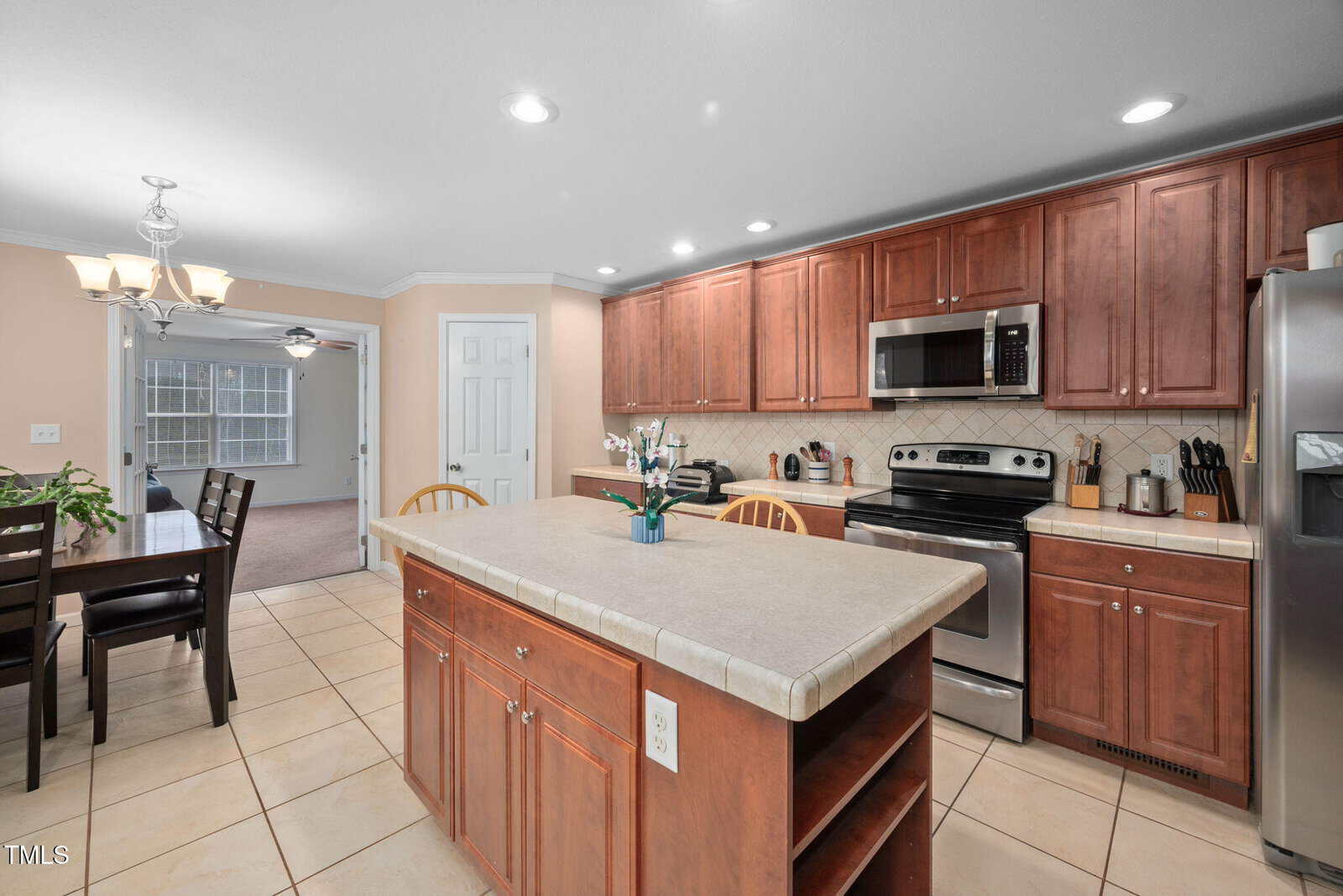 A kitchen with wooden cabinets and a tiled floor features an island in the center. Appliances include a stainless steel stove, microwave, and refrigerator. A dining table with chairs is visible in the adjoining room, which is carpeted.