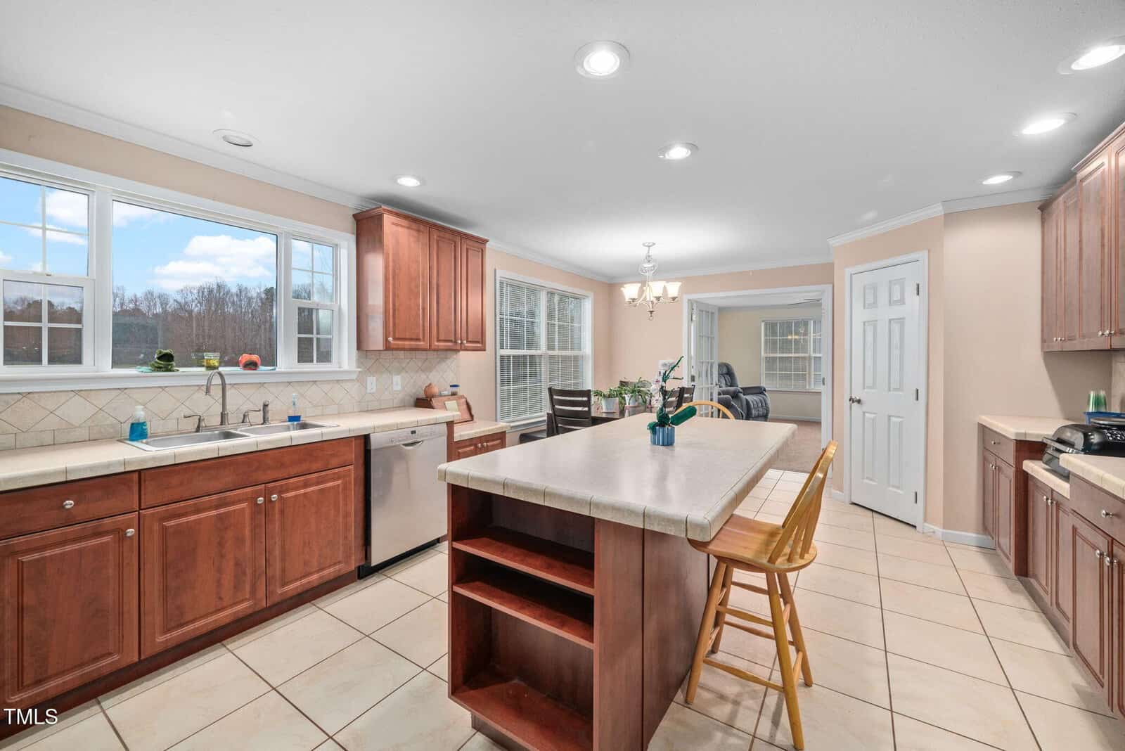 A spacious kitchen with wooden cabinets, a long island countertop with bar chairs, and a tiled floor. The kitchen has stainless steel appliances and large windows letting in natural light. A dining area is visible in the background.