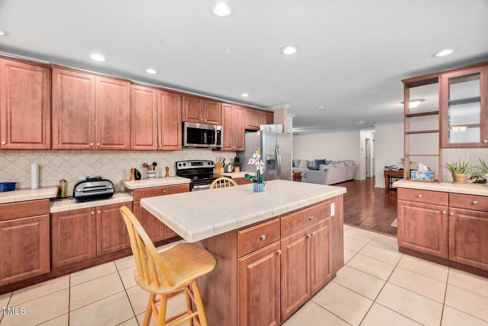Spacious kitchen with wooden cabinets and a tiled backsplash. An island with a beige countertop and wooden chair is central. Stainless steel appliances are included. Opening to a living area with hardwood flooring and a visible couch.