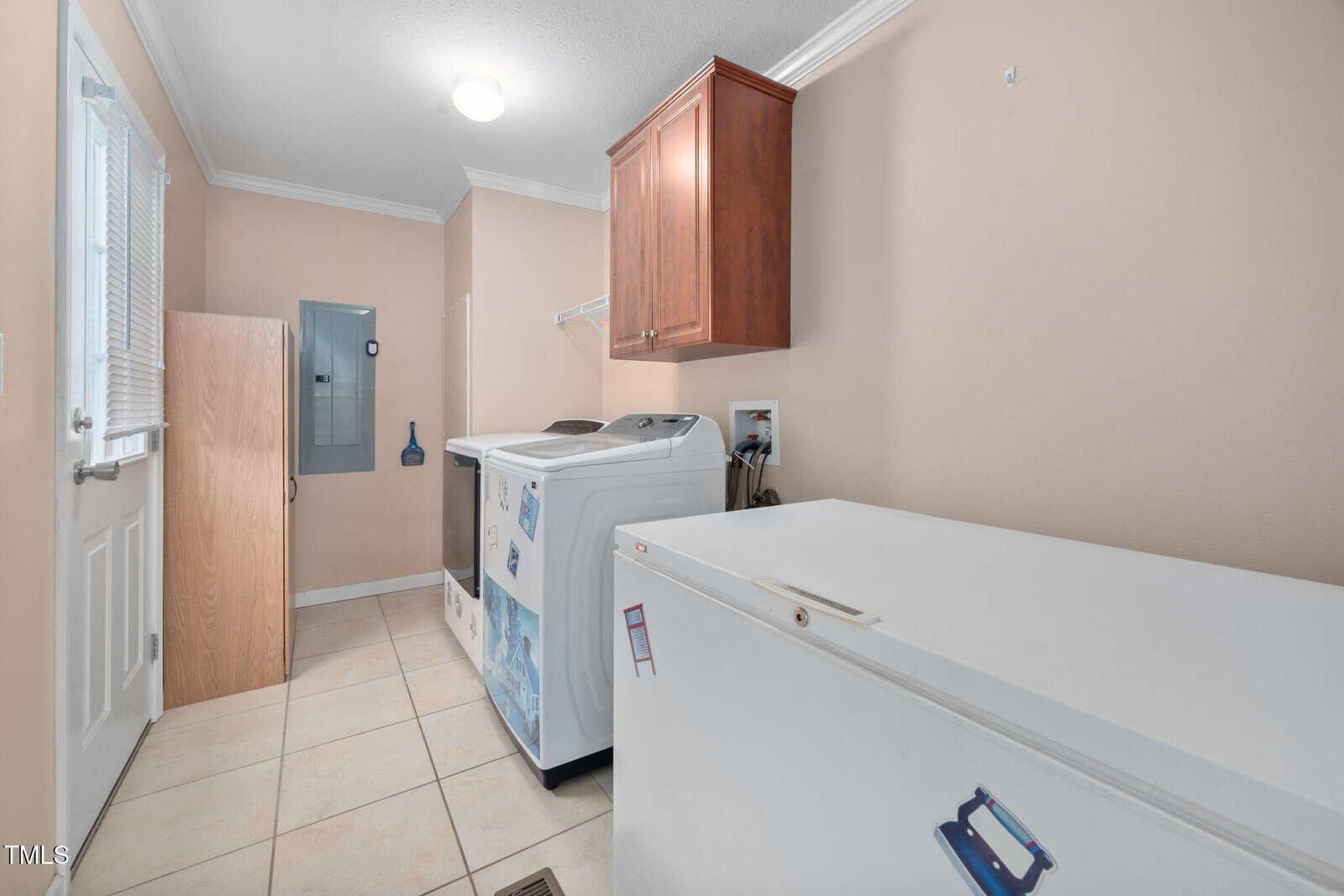A laundry room with beige walls and tiled flooring. It features a washing machine, dryer, and a white chest freezer. A wooden cabinet is mounted on the wall, and cleaning tools are stored in the corner. A small window is on the left.