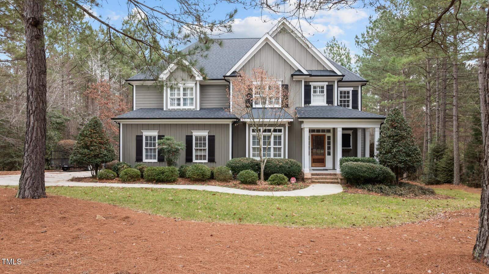 A two-story gray house with white trim and a covered front porch, surrounded by trees and shrubs, with a curved walkway leading to the front steps. Pinestraw covers the ground around the lawn.
