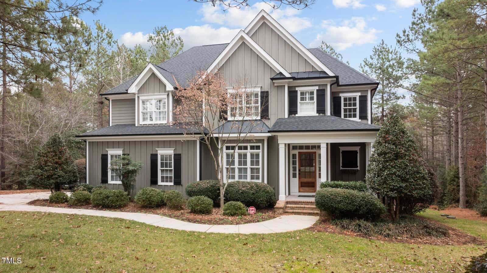 Two-story gray house with white trim, black shutters, and a covered front porch. The home is surrounded by bushes, trees, and a curved walkway. Pine trees and a partially cloudy sky are visible in the background.