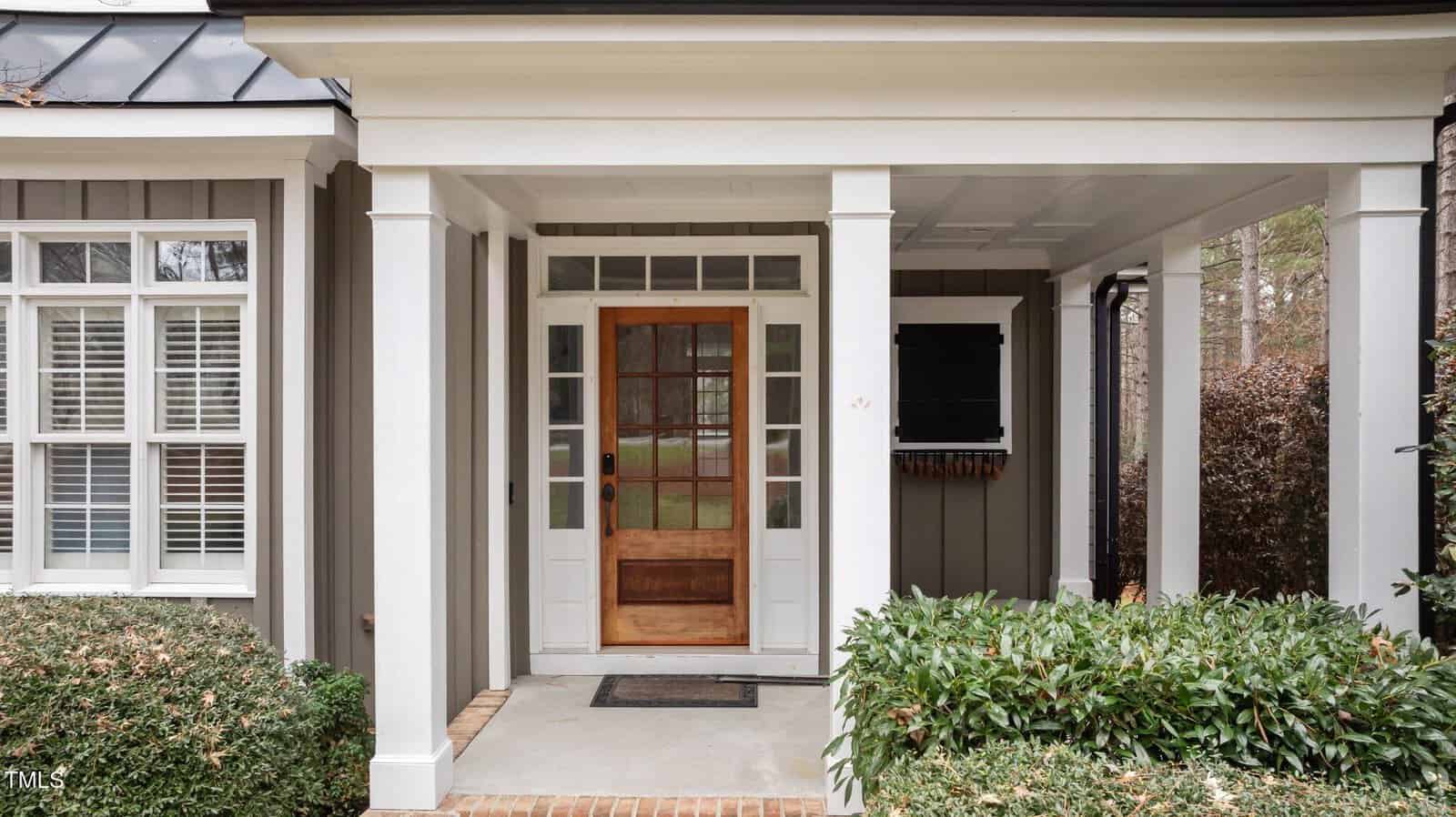 A wooden front door with glass panels is centered under a covered porch. White trim, square columns, two side windows, and shrubs surround the entrance. A black wall-mounted mailbox is to the right of the door.