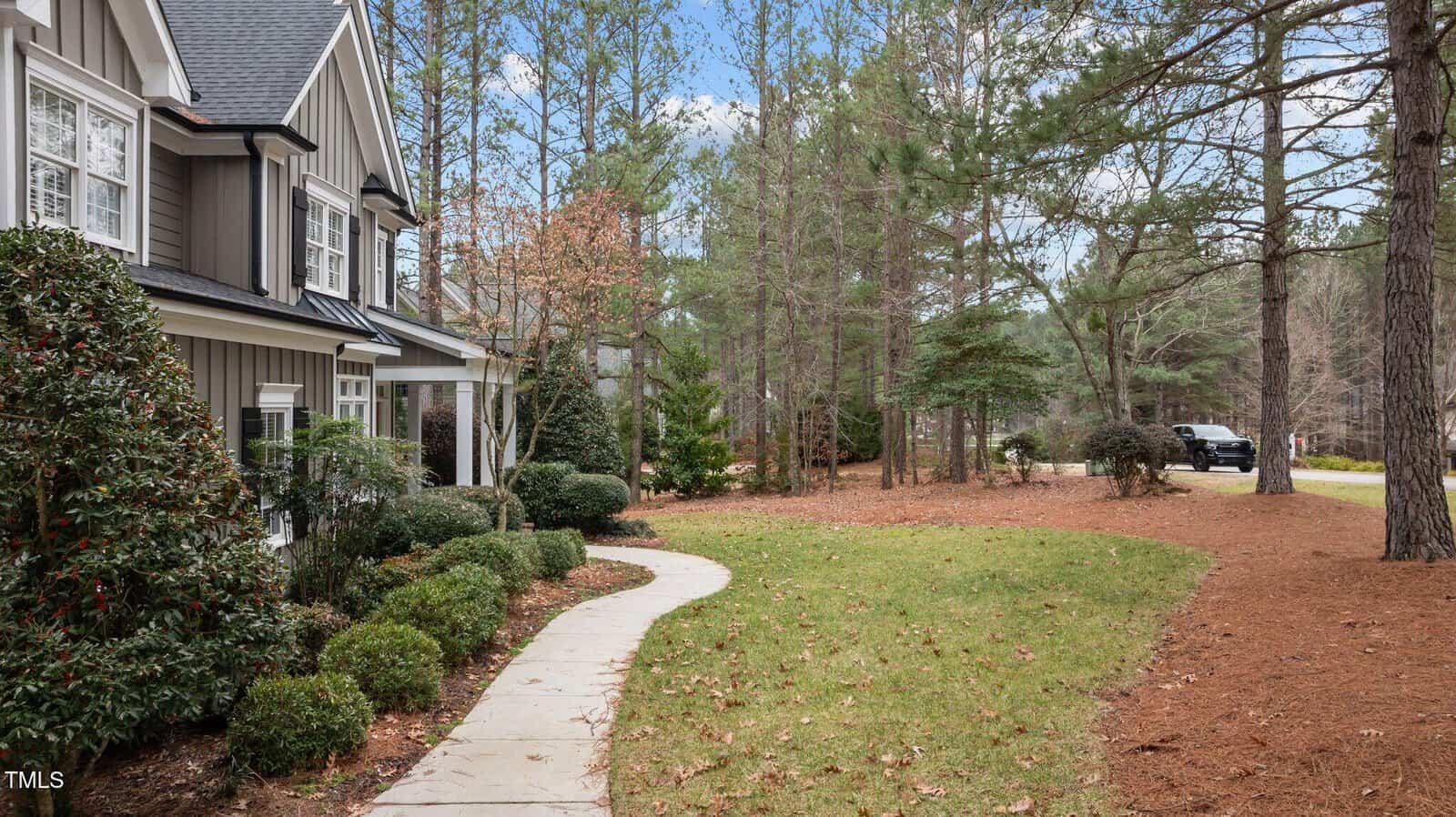 A gray house with white trim is on the left, bordered by bushes and a curved stone walkway. The yard is covered in pine straw with scattered leaves, and pine trees line the right side near a road with a parked black vehicle.