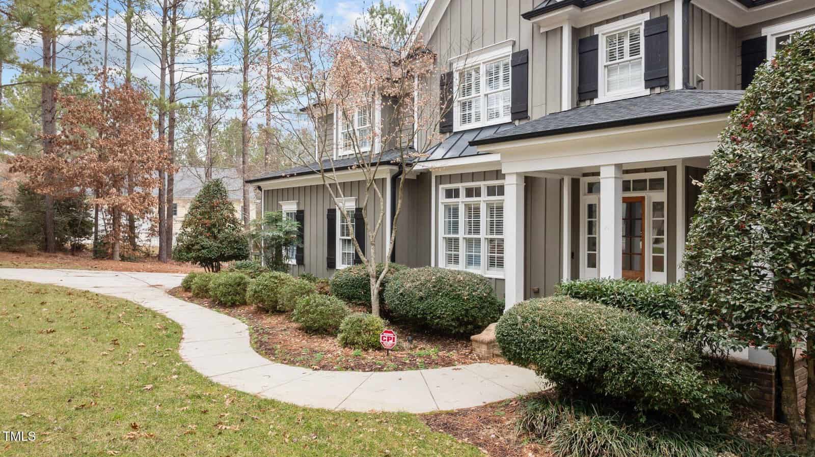 A curved concrete walkway leads to the front porch of a two-story house with gray siding, black shutters, and white trim. Bushes and trees are landscaped along the walkway and the porch.