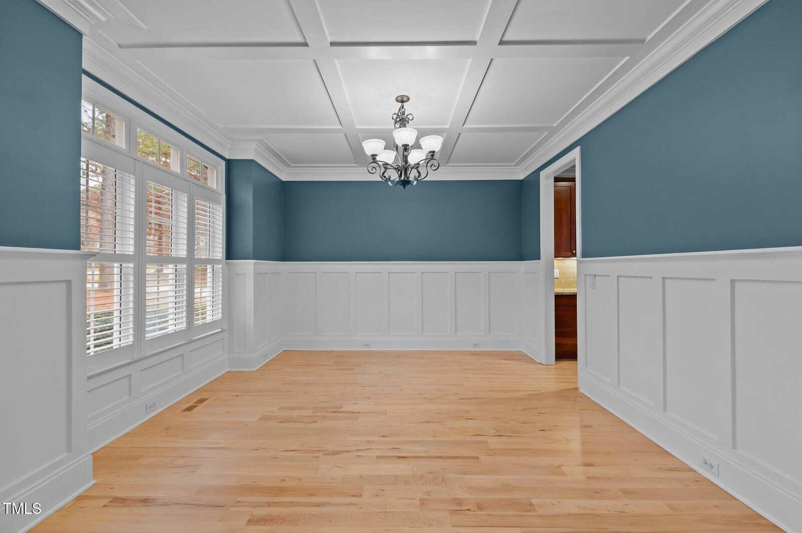 Empty dining room with blue upper walls, white wainscoting, light wood floors, a decorative chandelier, and large windows with white shutters. An open doorway leads to another room with wood cabinetry.