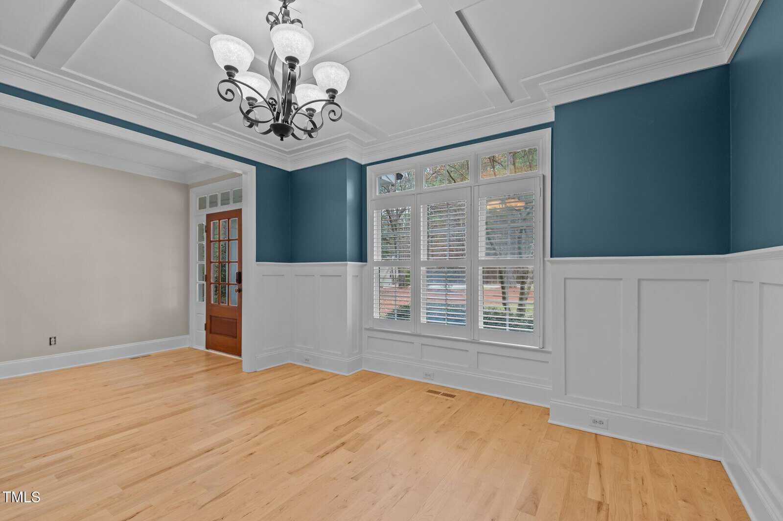 Empty dining room with light wood floors, white wainscoting, blue upper walls, a coffered ceiling, and a black chandelier. Large windows with blinds let in daylight, and a wooden door with glass panels is visible.