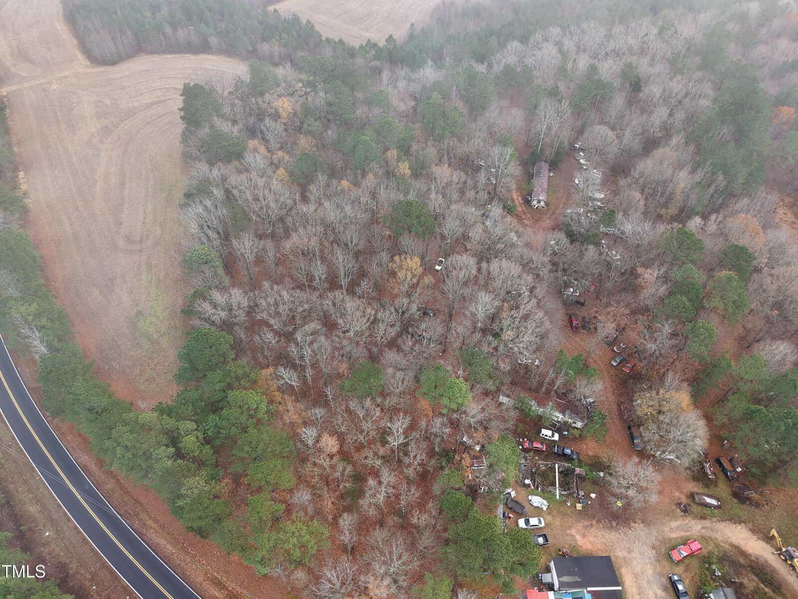 Aerial view of a wooded area near Louisburg with scattered vehicles and structures close to the edge, bordered by a curved road—0 Breedlove Road—on the left and an open field in the top left corner. Trees have sparse foliage, likely indicating autumn or winter.