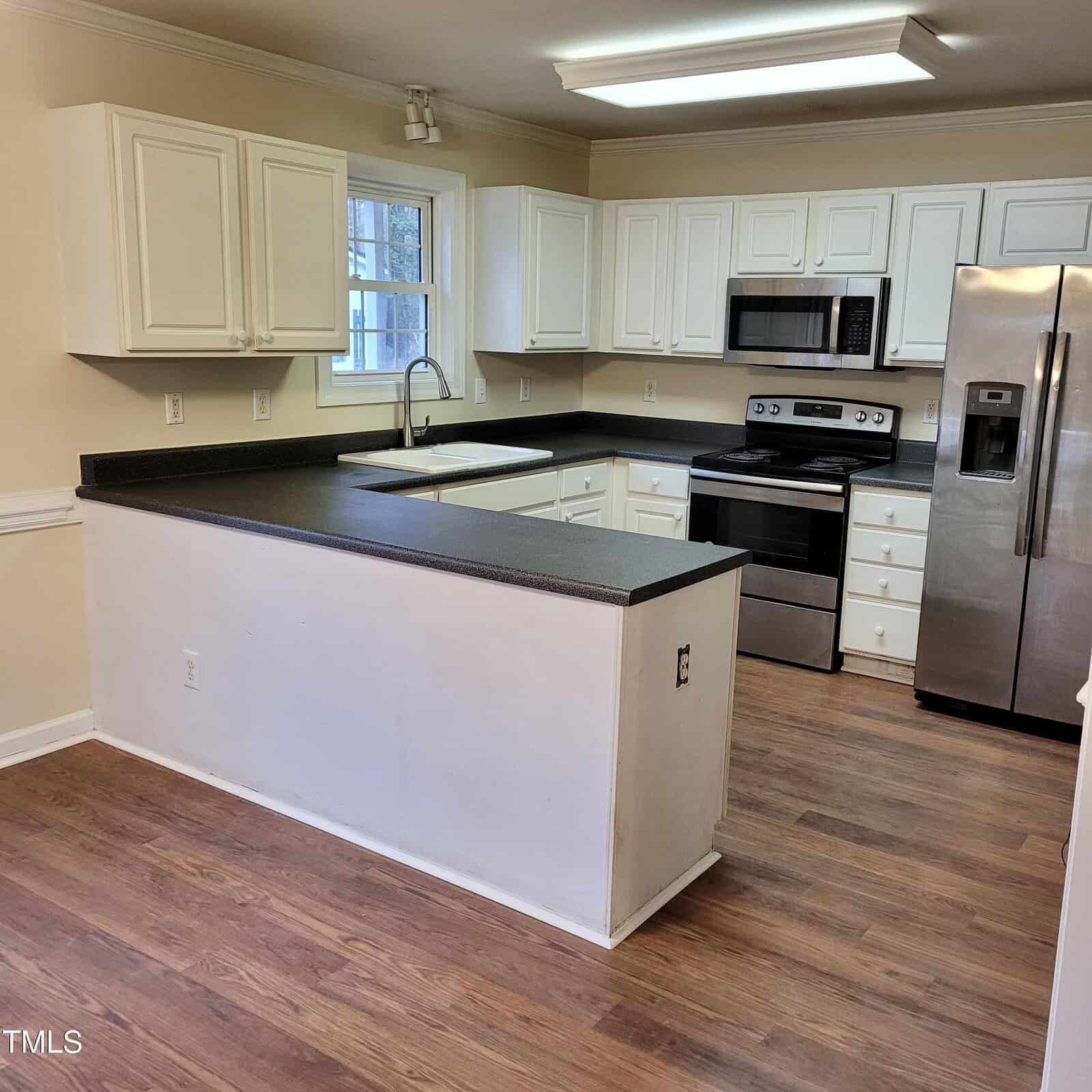A modern kitchen with white cabinets, black countertops, stainless steel appliances including a refrigerator, oven, and microwave, with wood flooring and a window above the sink.