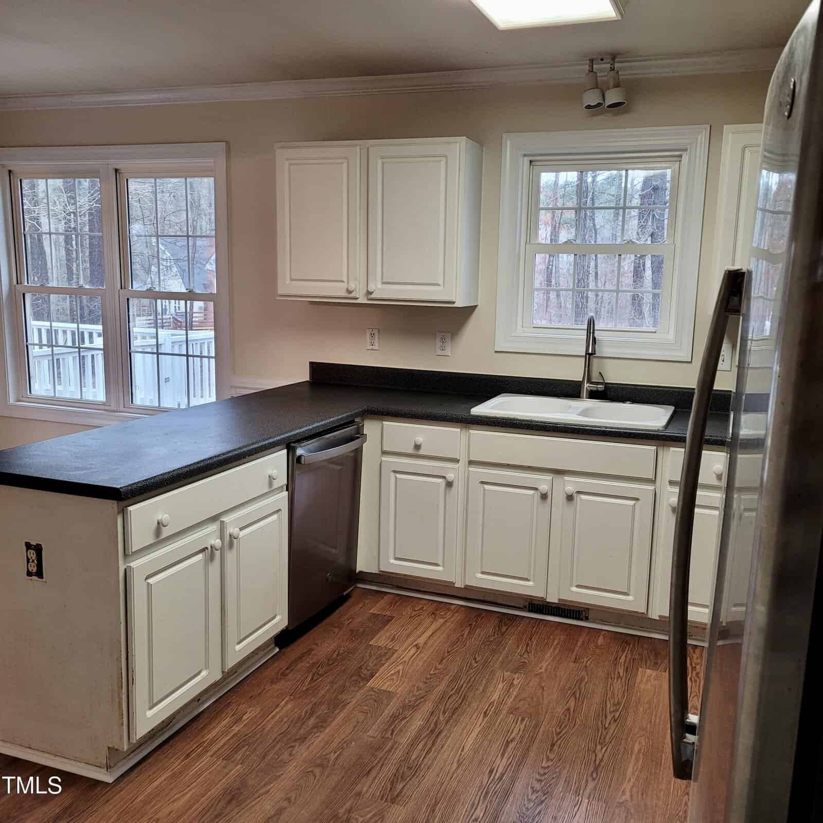 A kitchen with white cabinets, black countertops, a stainless steel dishwasher, a white sink under two windows, hardwood floors, and a refrigerator on the right. Walls are beige with no decorations.