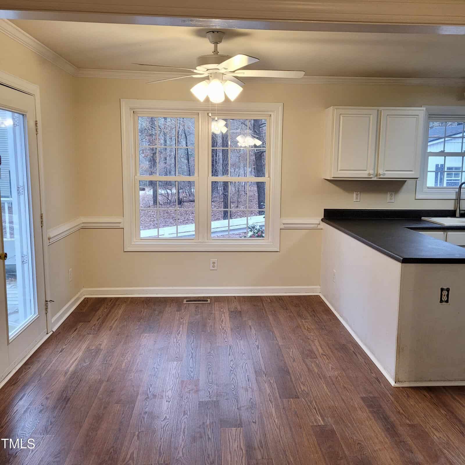 A room with wood floors, cream walls, a ceiling fan with lights, large window, partial black countertop with white cabinets, and a door leading outside. A wooded area is visible through the windows.